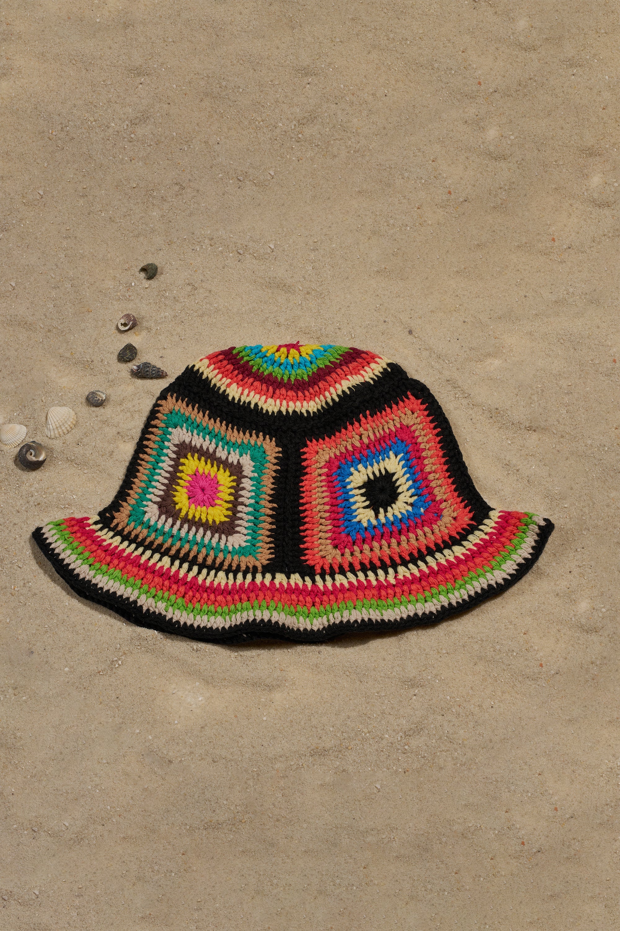 The DND CROCHET BUCKET HAT, featuring retro granny-square patterns, rests on sandy ground with a few seashells scattered around. 1