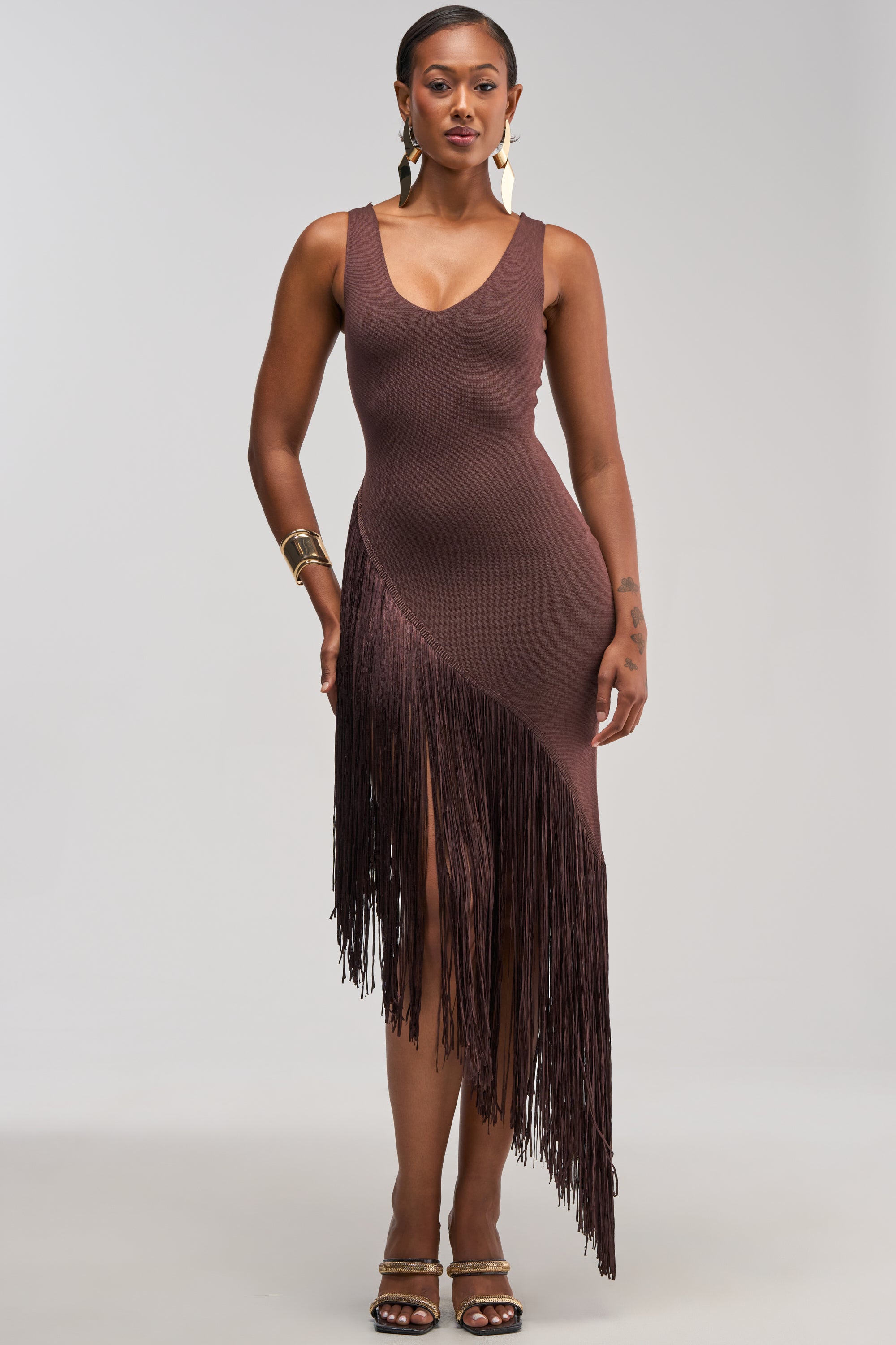 A woman stands against a plain light background wearing the SHAKE IT FRINGE MIDI DRESS IN BROWN, featuring an asymmetrical silhouette and sleeveless design, styled with gold earrings, bracelets, and heeled sandals.
