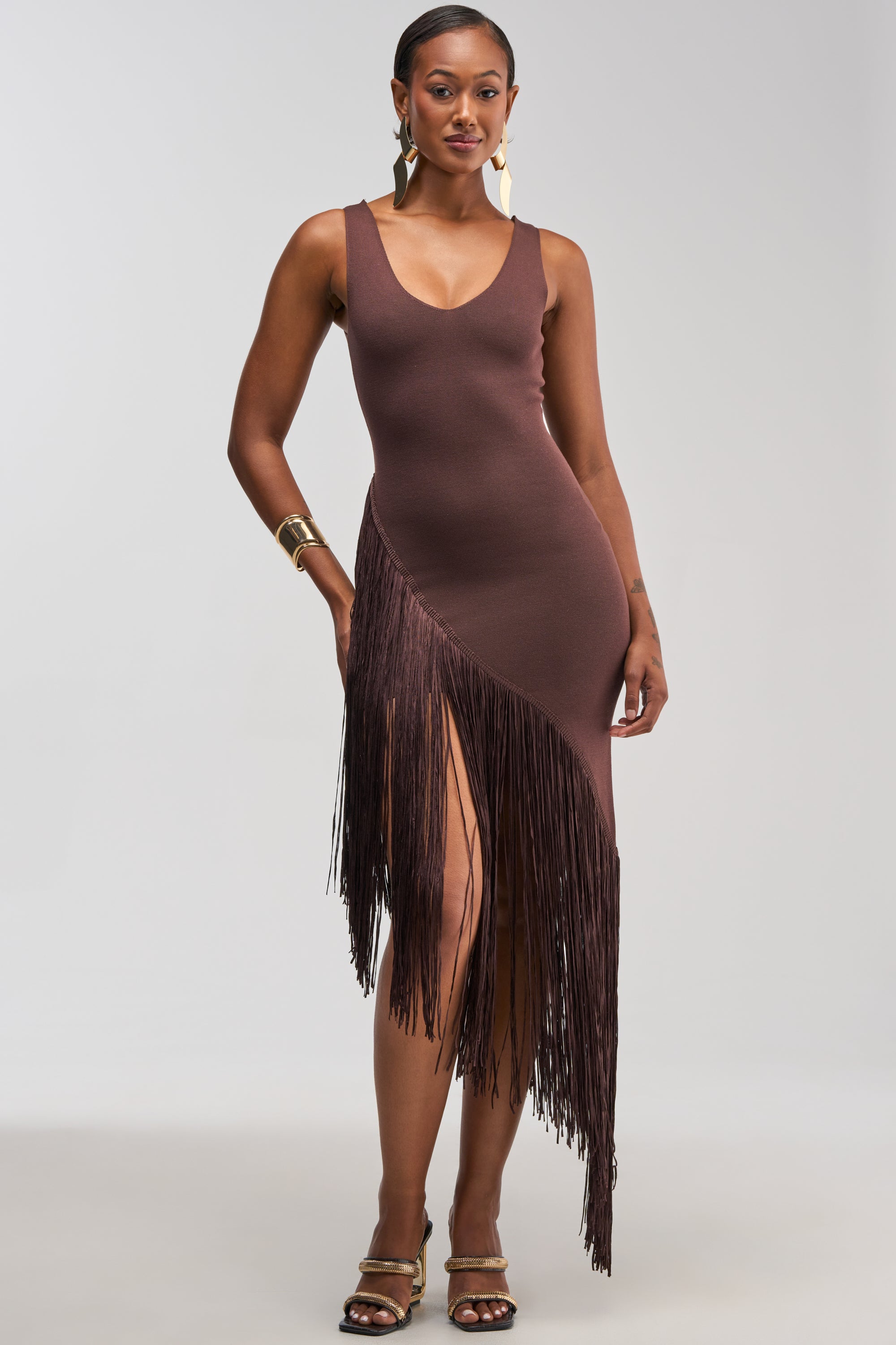 Wearing the SHAKE IT FRINGE MIDI DRESS IN BROWN, a woman stands confidently in this asymmetrical, sleeveless, form-fitting dress featuring a deep V-neckline and eye-catching fringe detail. Gold earrings, a bracelet, and sandals complete her look—making this the perfect cocktail party dress.