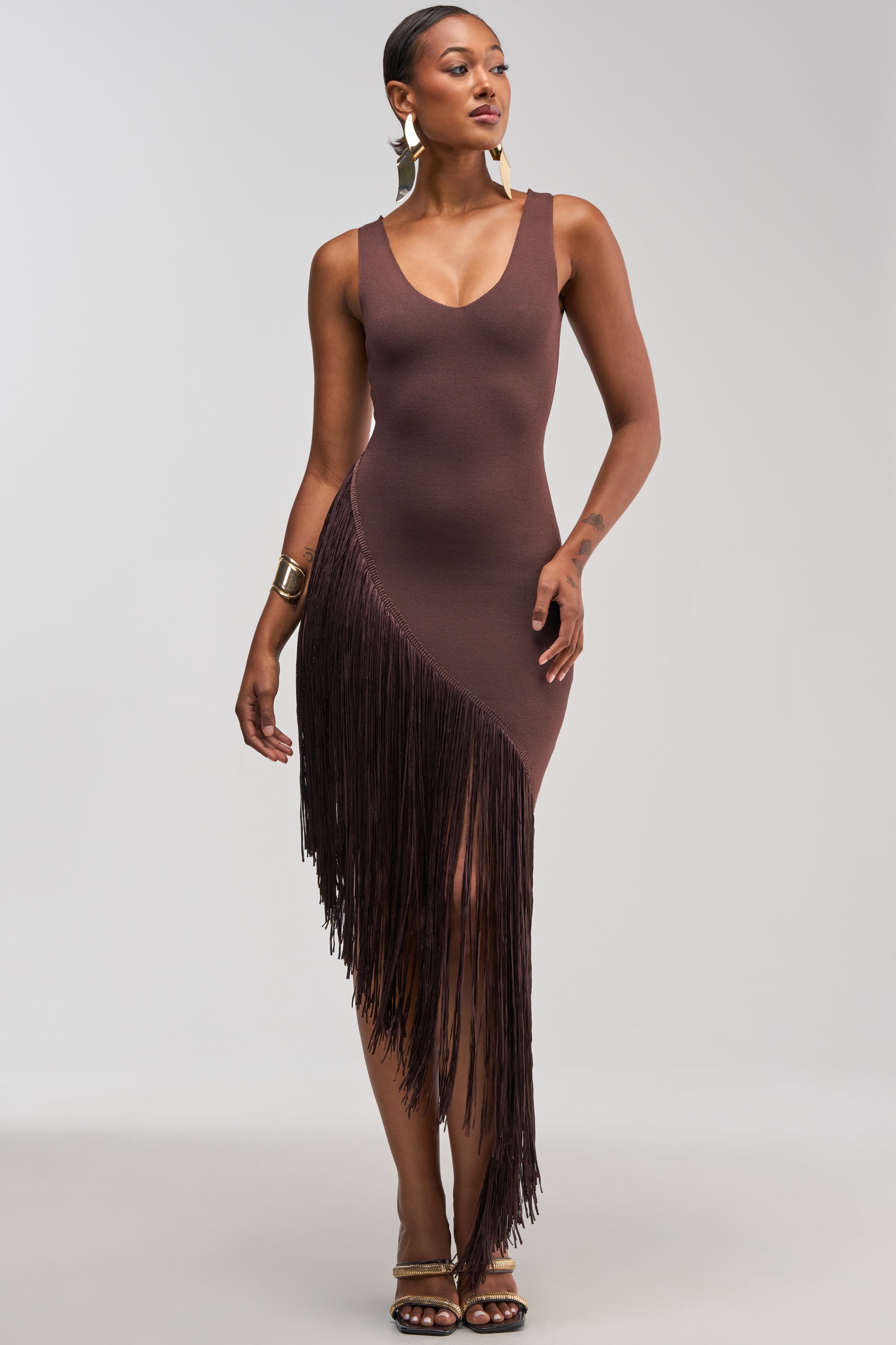 A woman wearing the SHAKE IT FRINGE MIDI DRESS IN BROWN—a sleeveless, fitted, asymmetrical brown dress with a fringed midi hem—styled with gold earrings and a gold bracelet, standing against a light background.