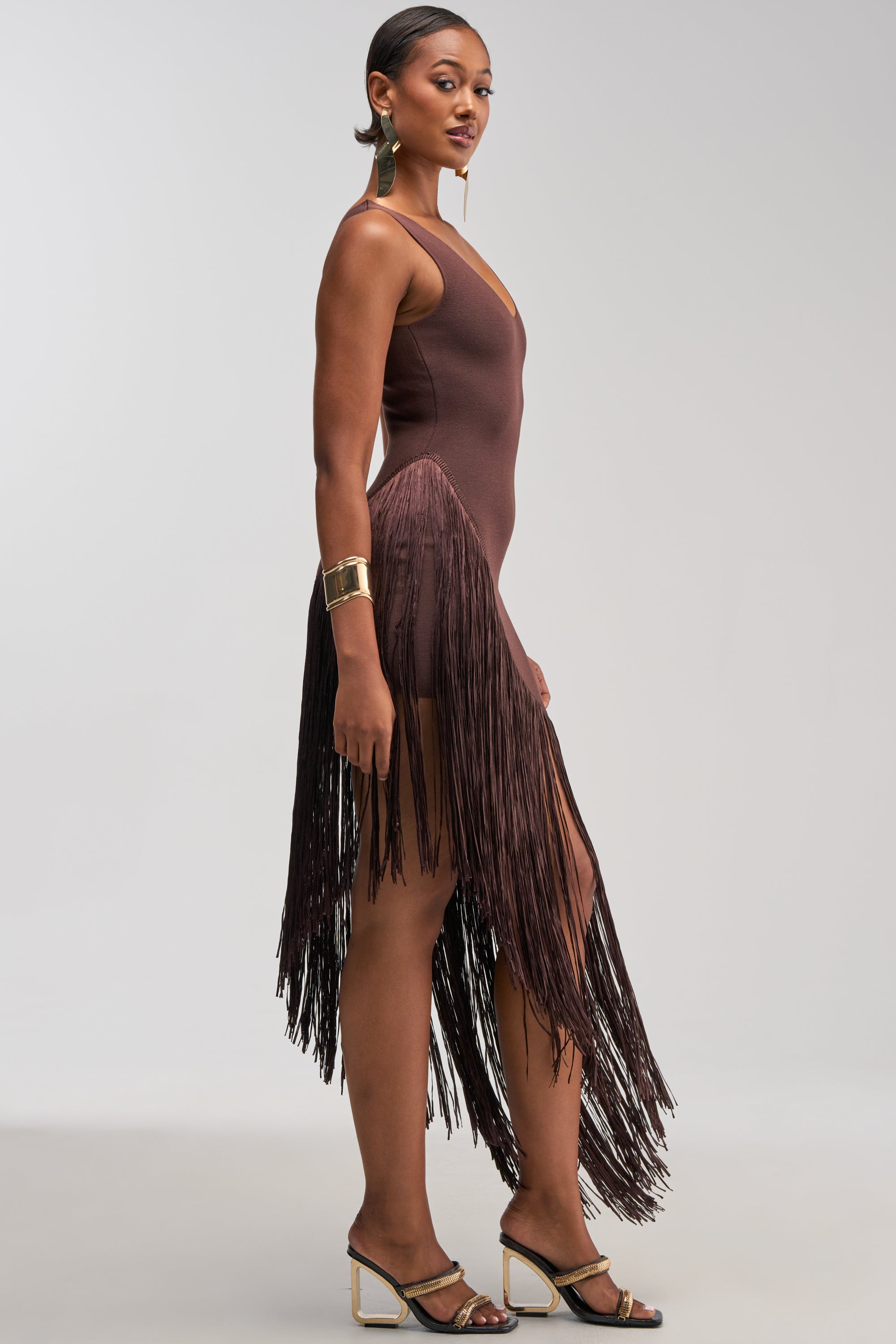 A woman in the SHAKE IT FRINGE MIDI DRESS IN BROWN stands in profile, styled with hoop earrings, a gold cuff bracelet, and strappy heels. Her hair is slicked back and her makeup is done.