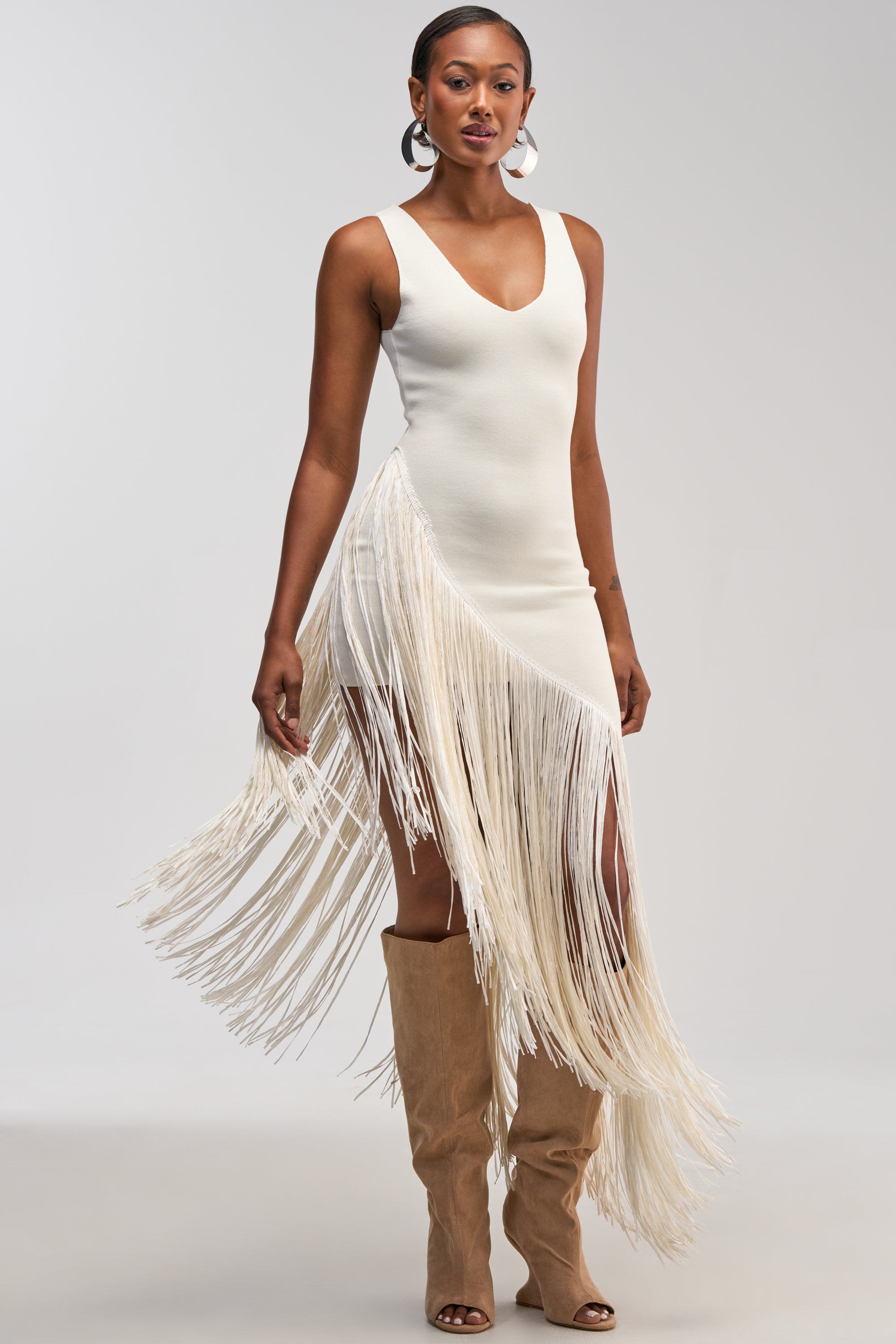 Wearing the SHAKE IT FRINGE MIDI DRESS IN WHITE—a sleeveless, asymmetrical midi dress featuring dramatic long fringe on one side—a woman stands against a light gray background, styled with tall tan suede boots and large hoop earrings.