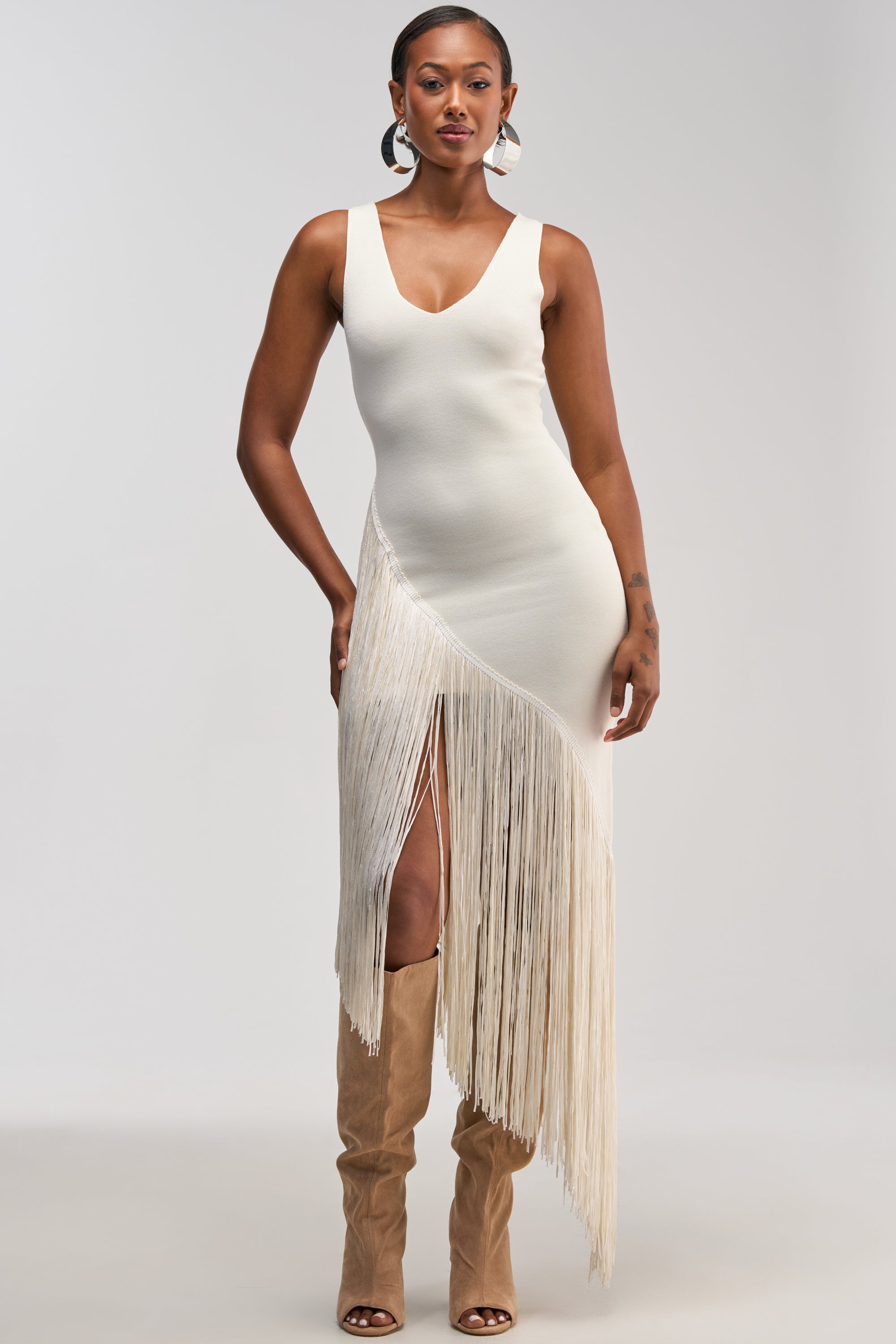 Dressed in the SHAKE IT FRINGE MIDI DRESS IN WHITE—a sleeveless, fitted white midi featuring bold fringe details and a dramatic asymmetrical hem—she pairs the look with tall tan boots. Ideal for cocktail parties or sophisticated bridal occasions, she exudes confidence against a plain backdrop.
