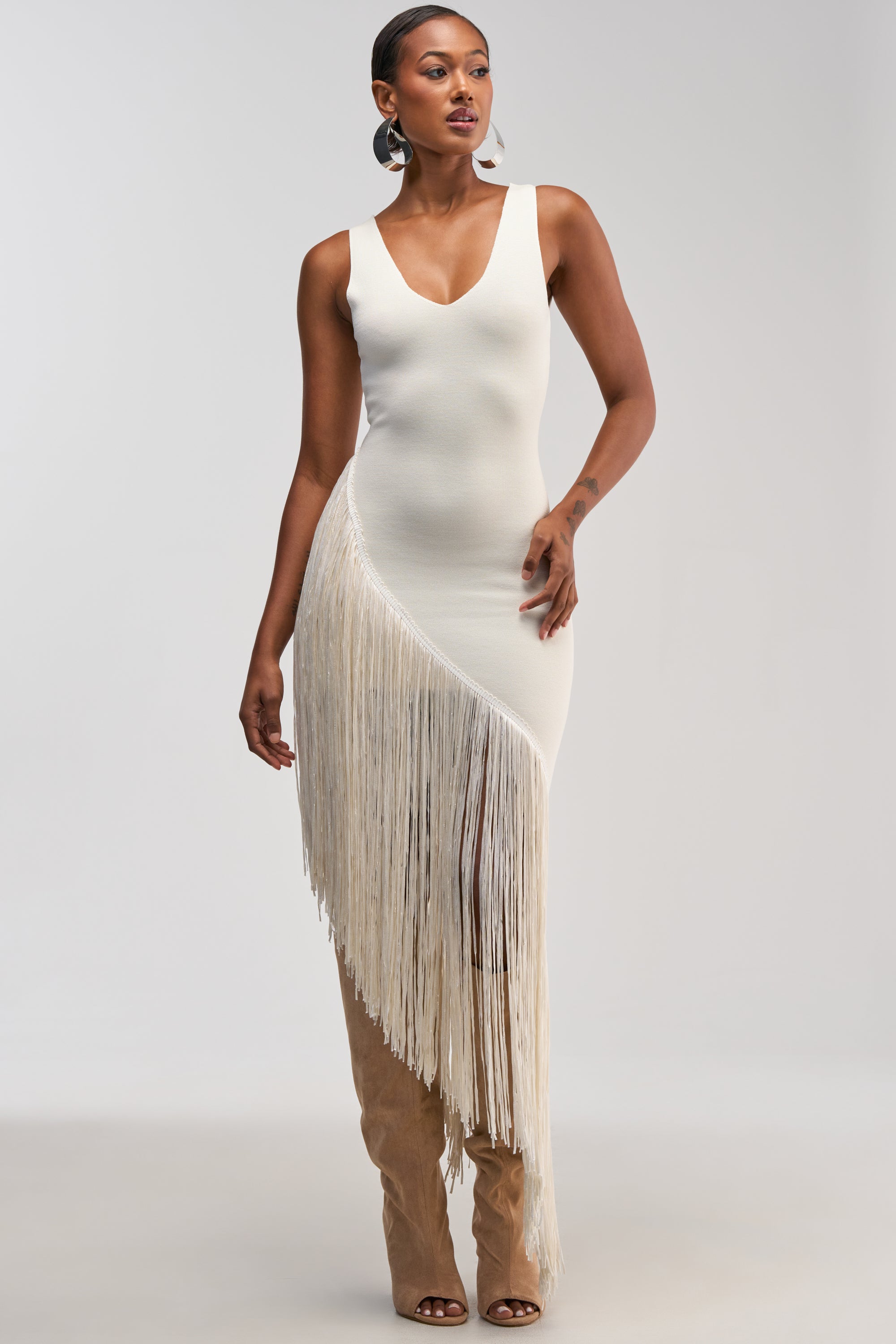 Model showcasing the SHAKE IT FRINGE MIDI DRESS IN WHITE—a sleeveless, fitted midi dress featuring an asymmetrical hem, deep V-neckline, and dramatic long fringe on one side—styled with tall tan boots and large hoop earrings against a plain background. An ideal choice for bridal events or a chic cocktail party look.