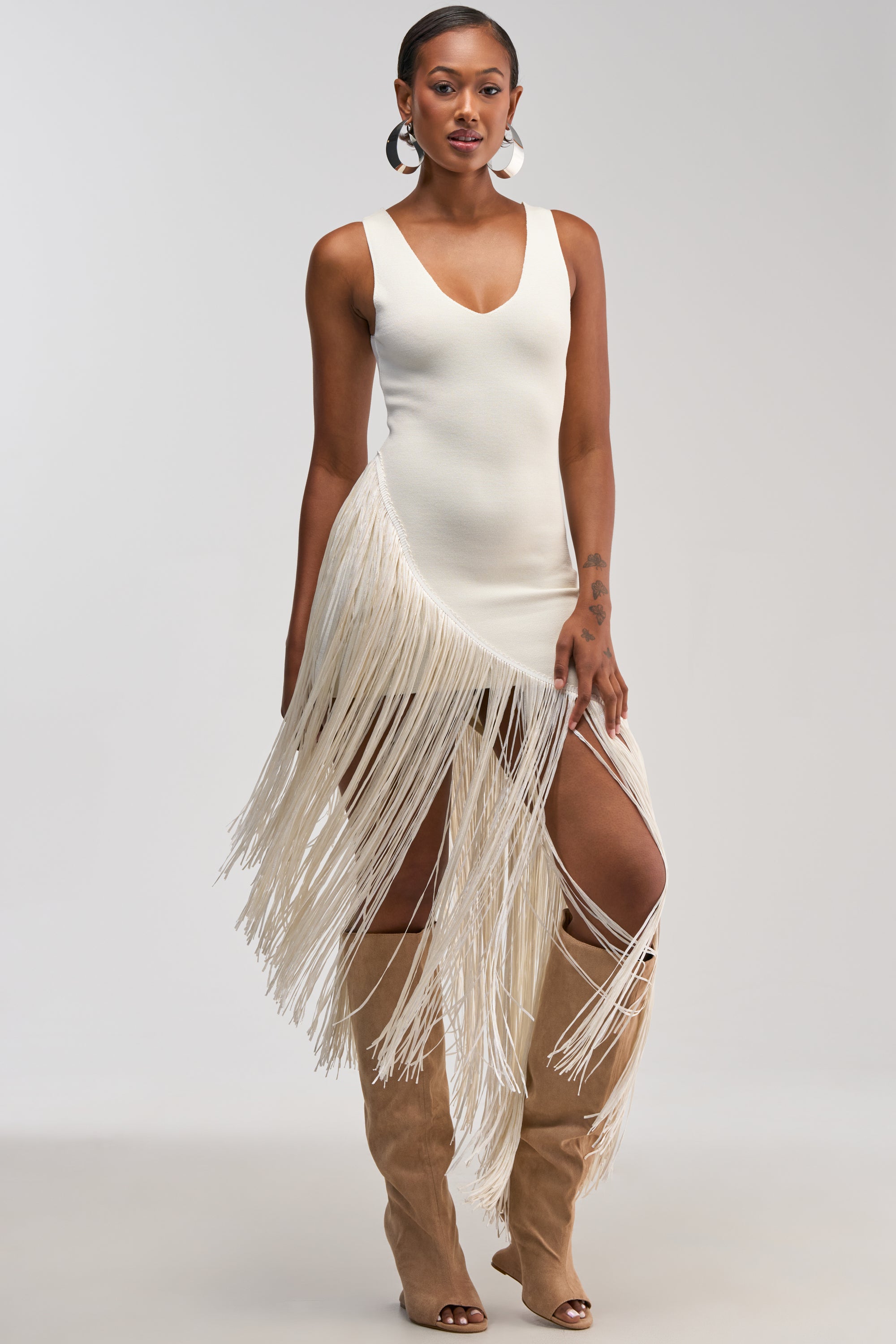 Woman wearing the SHAKE IT FRINGE MIDI DRESS IN WHITE, featuring an asymmetrical silhouette with a sleeveless white bodice and long fringes cascading down one side. She pairs it with tan knee-high boots and stands against a plain light background—an ideal look for cocktail parties or bridal event occasions.