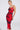 A woman models the ROSEY MIDI DRESS—a red, sleeveless bodycon dress with black abstract patterns, thin straps, and an open-back—standing confidently against a white background with slicked-back hair and large hoop earrings.