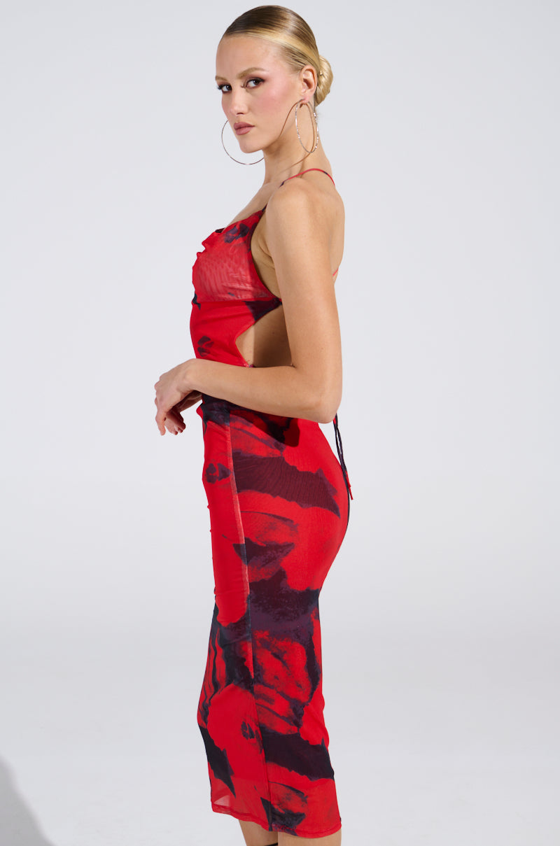 A woman poses in side profile wearing the ROSEY MIDI DRESS, a curve-hugging, sleeveless red and black floral dress with thin straps. Her hair is styled in a sleek bun, accessorized with large hoop earrings against a plain light background.
