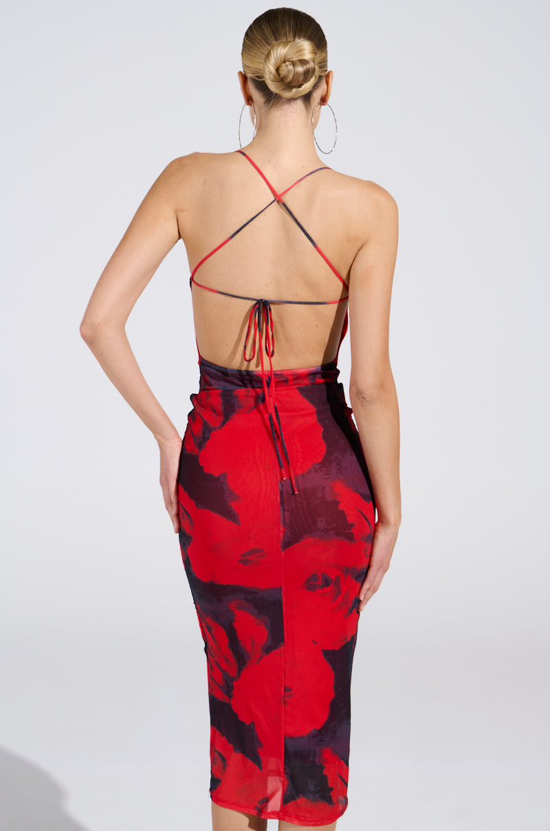 A woman with blonde hair in a sleek bun wears the ROSEY MIDI DRESS—a red and black floral, curve-hugging, backless halter-neck dress with crisscross straps—standing against a plain light background.