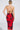 A woman with blonde hair in a sleek bun wears the ROSEY MIDI DRESS—a red and black floral, curve-hugging, backless halter-neck dress with crisscross straps—standing against a plain light background.