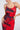 A woman poses confidently with one hand on her hip, wearing the ROSEY MIDI DRESS, a red and black tie-dye sleeveless dress featuring thin straps and an open-back design. She accessorizes with large hoop earrings against a light background.