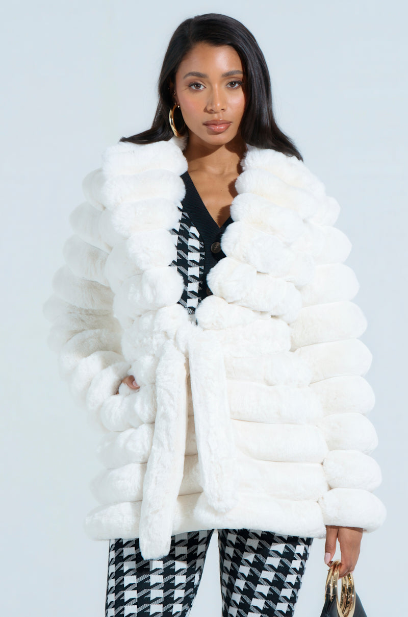 A woman poses confidently in a luxurious LANA POPCORN FAUX FUR JACKET, featuring an oversized silhouette and self-tie waist belt. Her long dark hair and gold hoop earrings complement the chic black and white houndstooth outfit underneath.