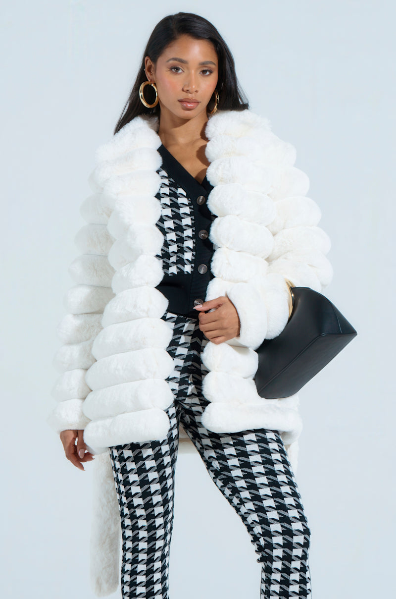 The woman wears a LANA POPCORN FAUX FUR JACKET, paired with a black top featuring bold buttons. Her black and white patterned pants complement the chic ensemble while she carries a sleek black bag. Gold hoop earrings accentuate her long hair against the simple light background.