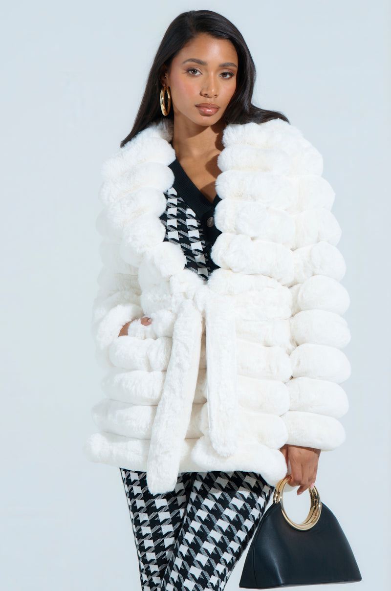 A woman confidently poses in a LANA POPCORN FAUX FUR JACKET with an oversized silhouette. She has long dark hair, wears black and white patterned pants, gold hoop earrings, and holds a black handbag featuring a gold handle against a plain background.