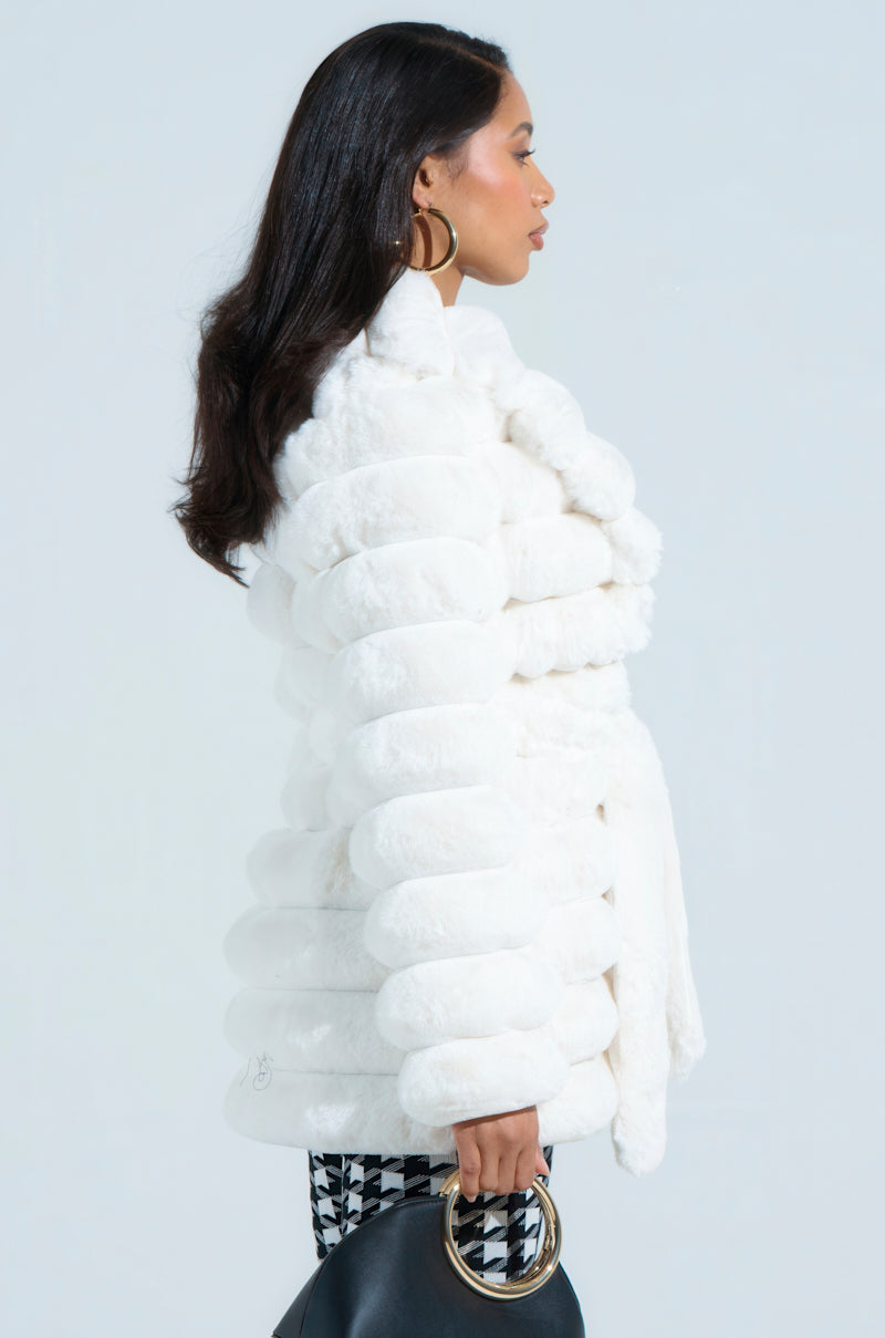 A woman in profile models the LANA POPCORN FAUX FUR JACKET, a plush white faux fur piece with an oversized silhouette. Her long dark hair and hoop earrings add style, while she holds a black handbag with gold handles and wears patterned pants for a chic touch.