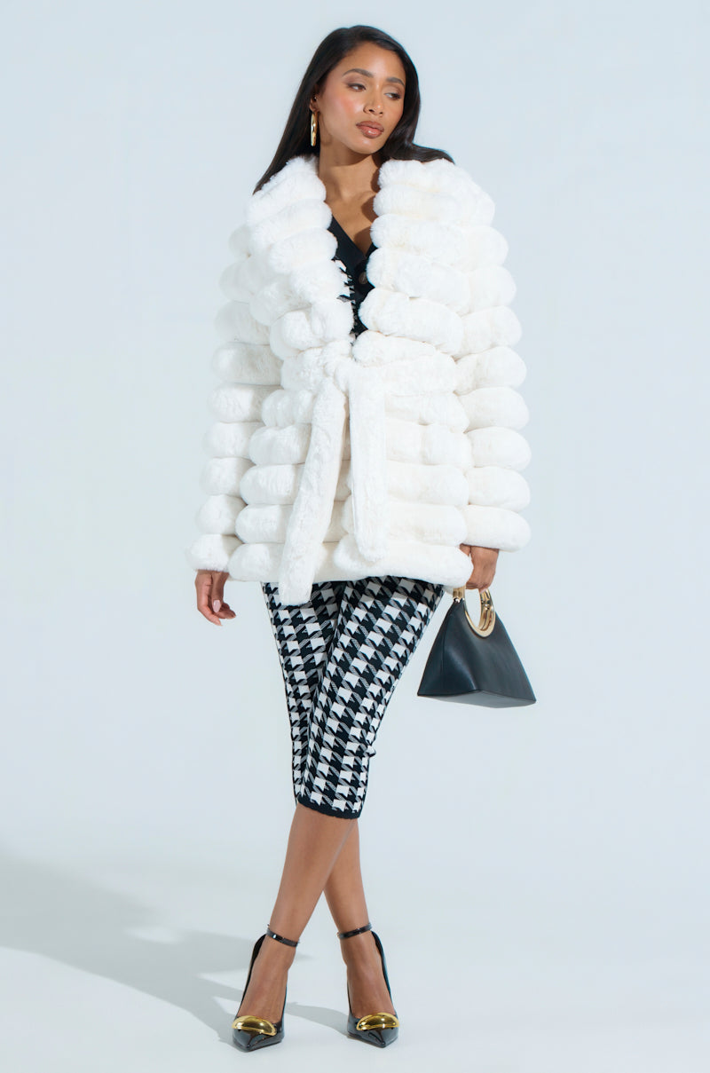 A woman wears the oversized LANA POPCORN FAUX FUR JACKET in white with black and white houndstooth pants and black heels featuring gold accents. She holds a small black purse, all against a plain light gray background.