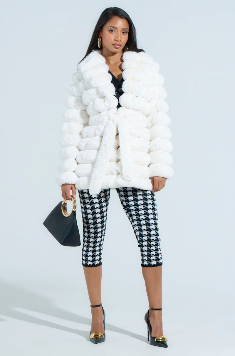 A person with long dark hair stands against a plain background wearing the LANA POPCORN FAUX FUR JACKET, black and white checkered capri pants, and black high-heeled shoes, holding a black handbag.