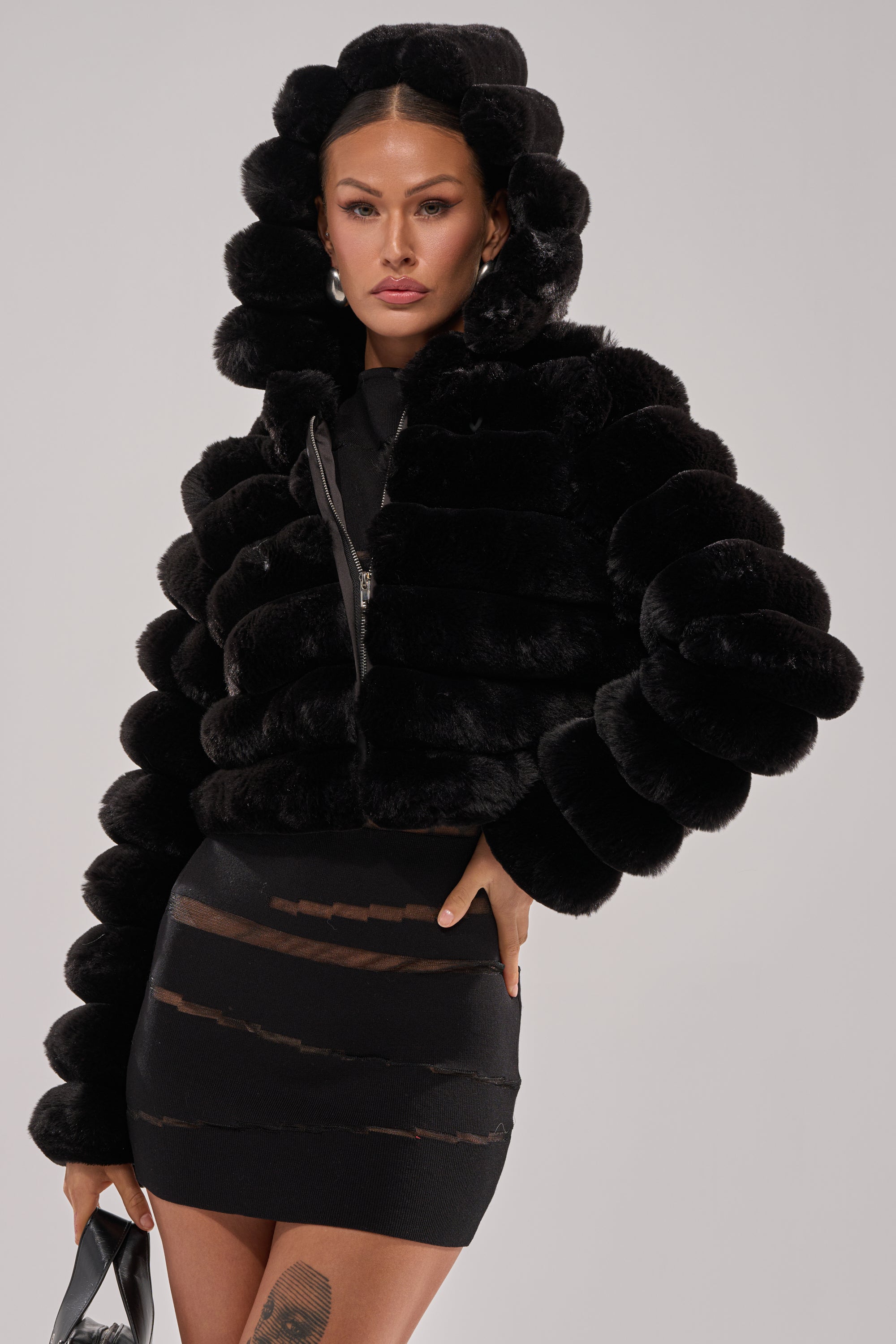 Woman wearing the POPCORN FAUX FUR IN BLACK jacket with a hood, paired with a sheer black mini skirt and holding a handbag, posing against a plain light background.