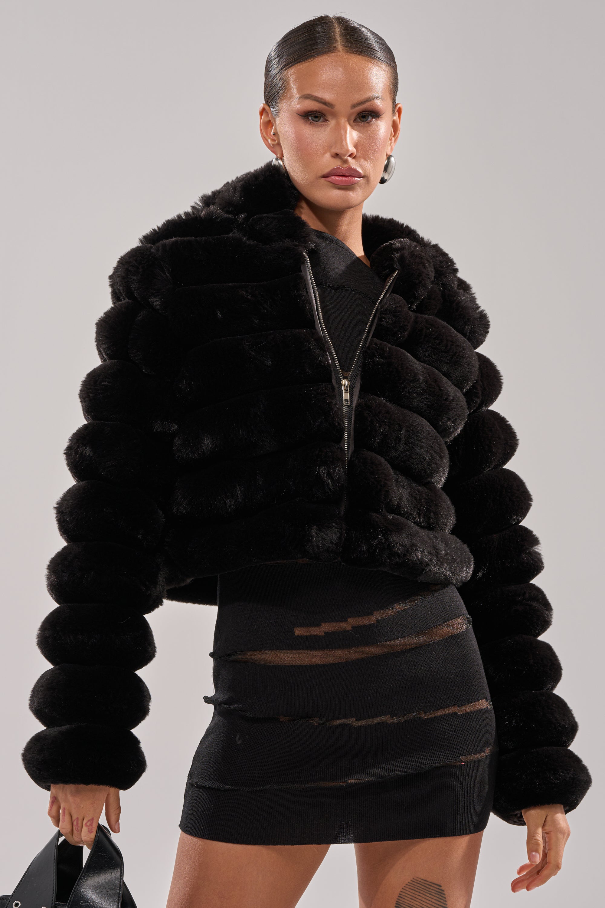 A woman wears the POPCORN FAUX FUR IN BLACK over a fitted black mini dress with sheer panels. She holds a black bag, her hair slicked back, posing against a plain light background.