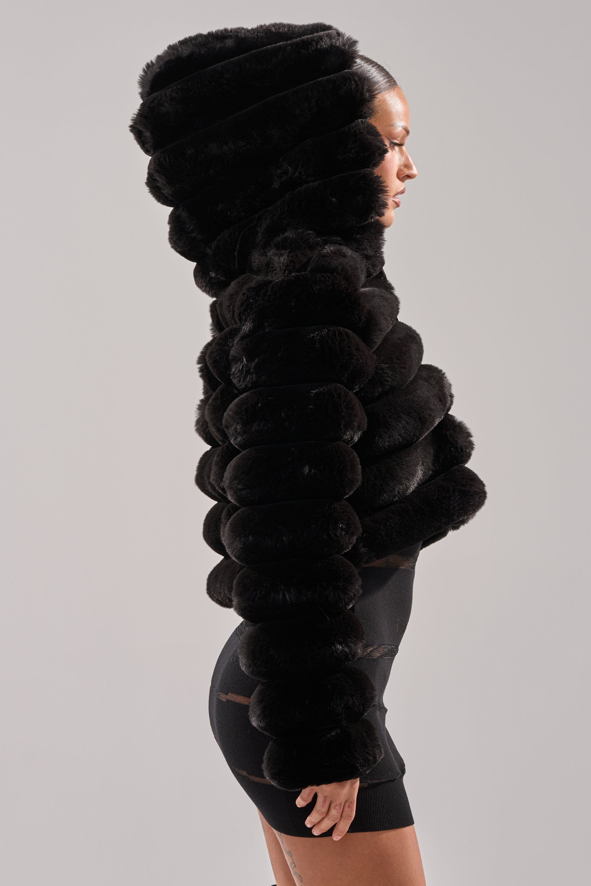 A woman in profile models the POPCORN FAUX FUR IN BLACK jacket, featuring a sculpted, puffy silhouette and large hood, paired with a fitted black dress, set against a plain light gray background.