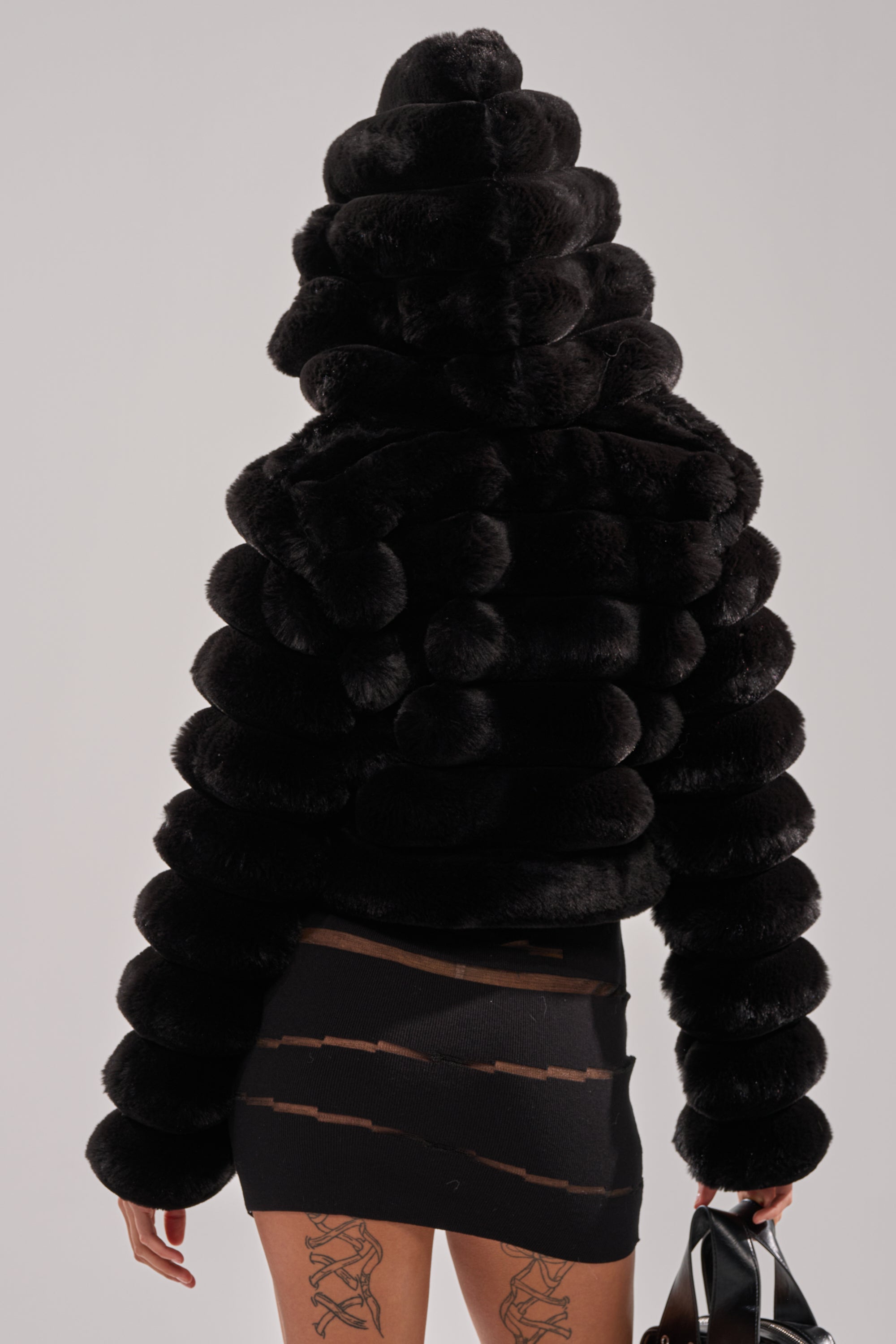 Wearing the POPCORN FAUX FUR IN BLACK, styled with a black mini skirt with sheer horizontal slits and a black handbag, against a plain background.