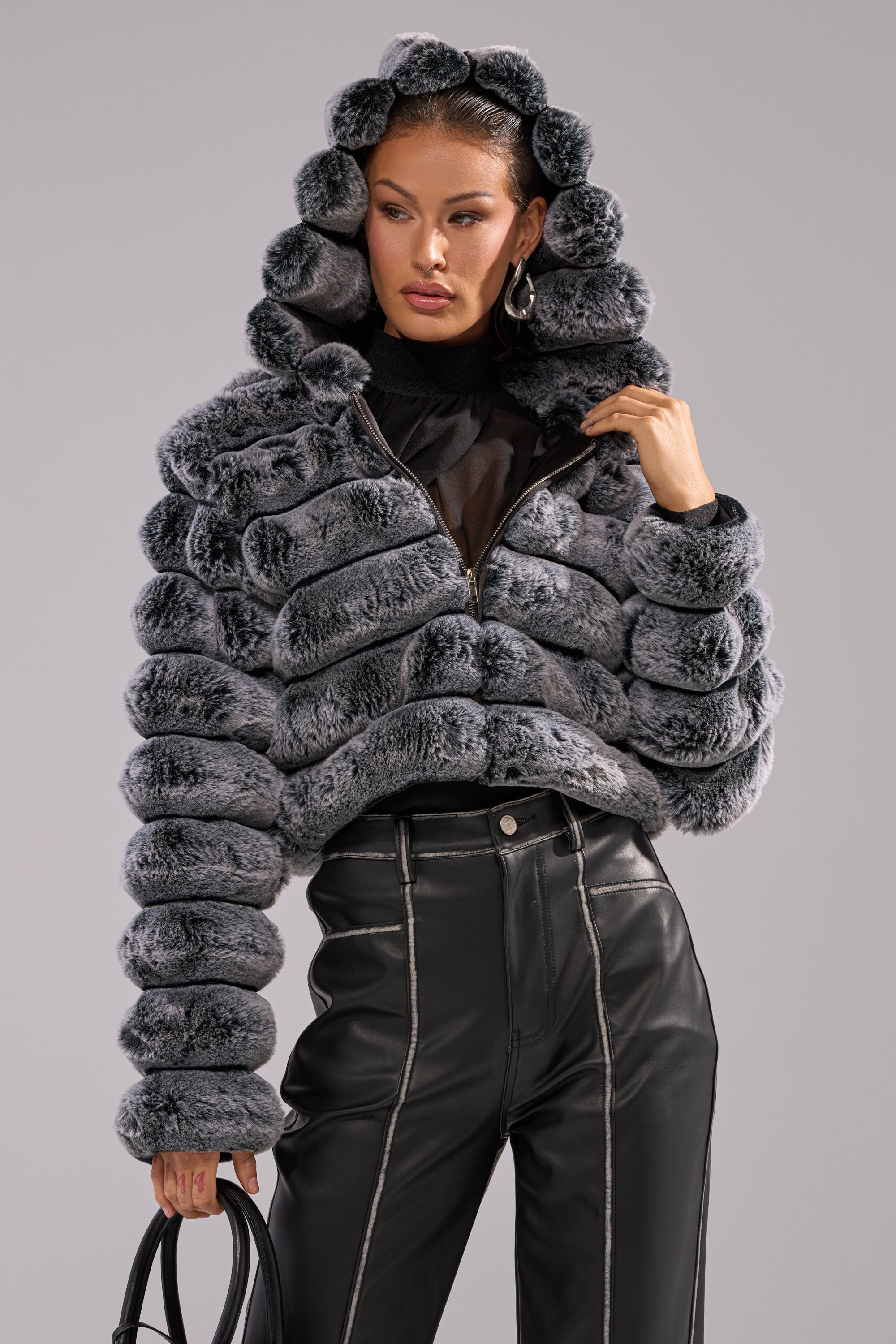 A woman stands confidently against a neutral background, wearing the POPCORN FAUX FUR IN GREY jacket with high-waisted black leather pants and holding a black handbag. 0