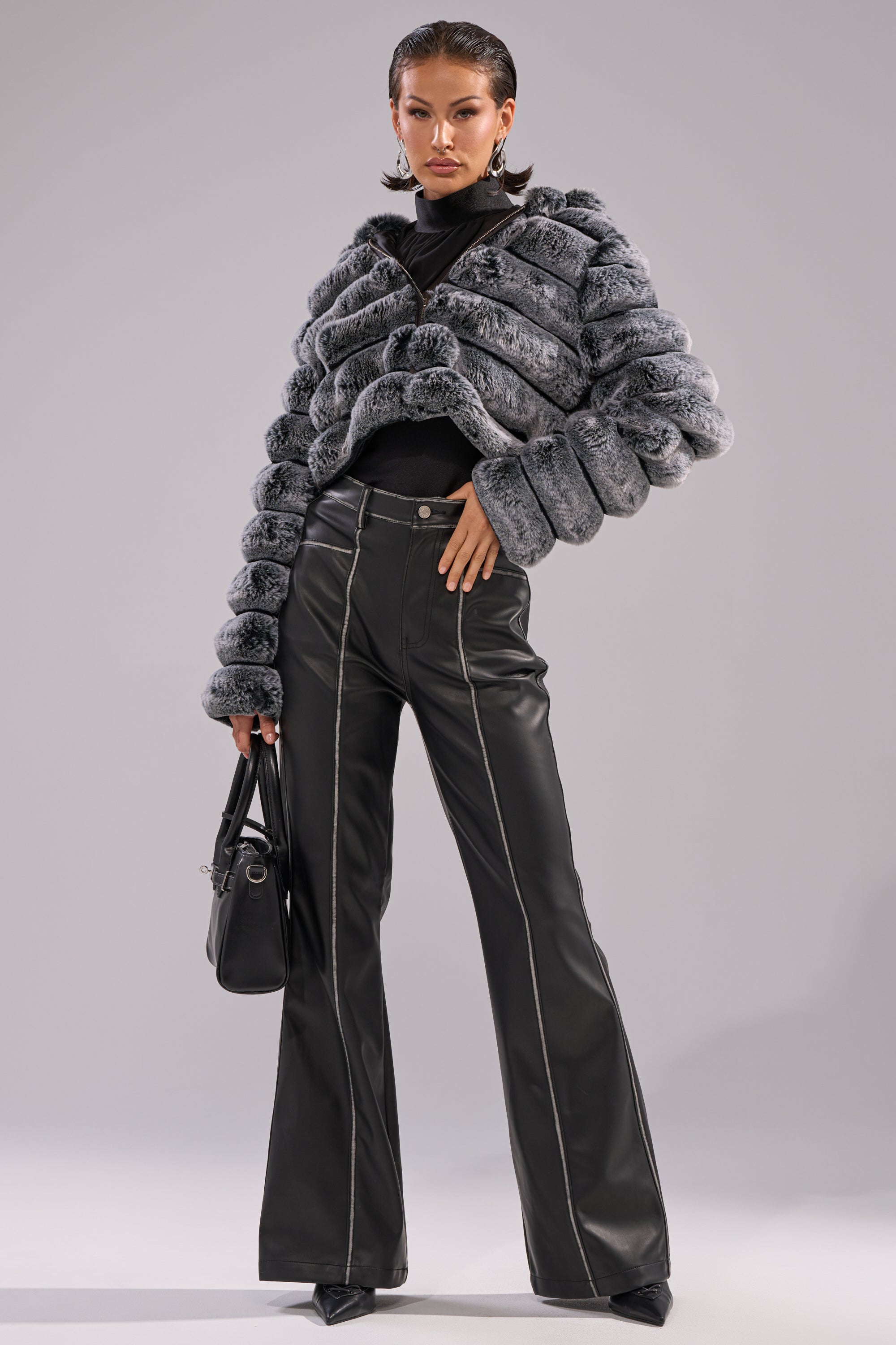 A woman poses confidently in the POPCORN FAUX FUR IN GREY jacket over a black turtleneck and high-waisted black leather pants with white stitching, accessorized with pointed heels and a black handbag against a plain gray background. 9
