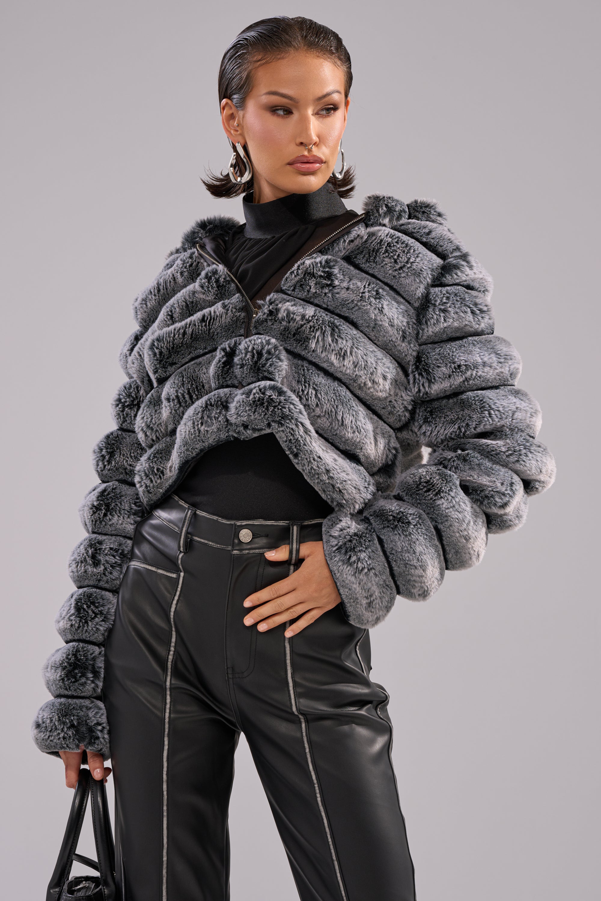 A woman models the POPCORN FAUX FUR IN GREY jacket over a black top and high-waisted black leather pants with white stitching, accessorized with a black handbag, posing confidently against a neutral background. 1
