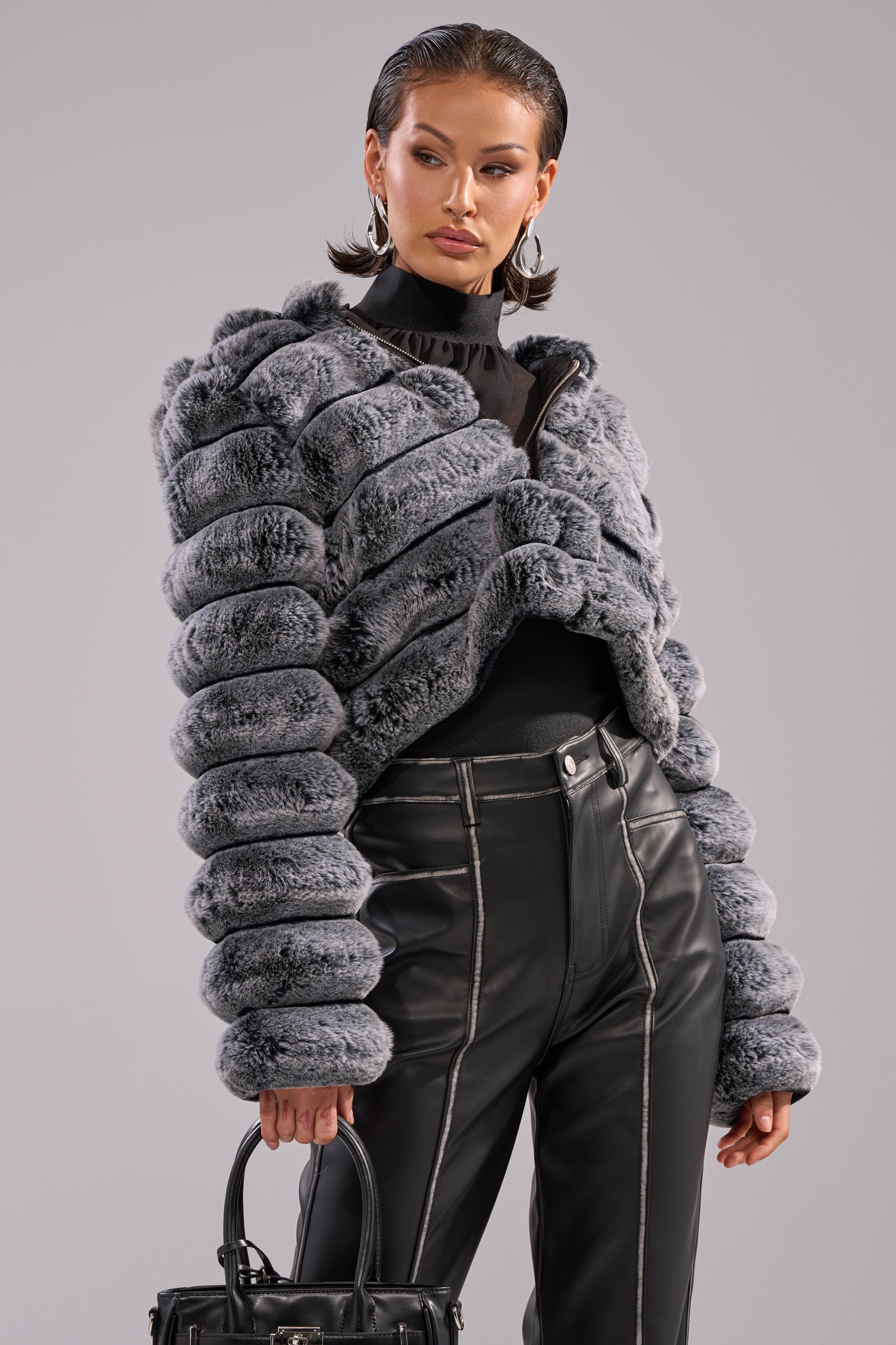 A woman showcases the POPCORN FAUX FUR IN GREY jacket, styled with a black turtleneck, high-waisted black leather pants, and a black handbag against a neutral background. 2