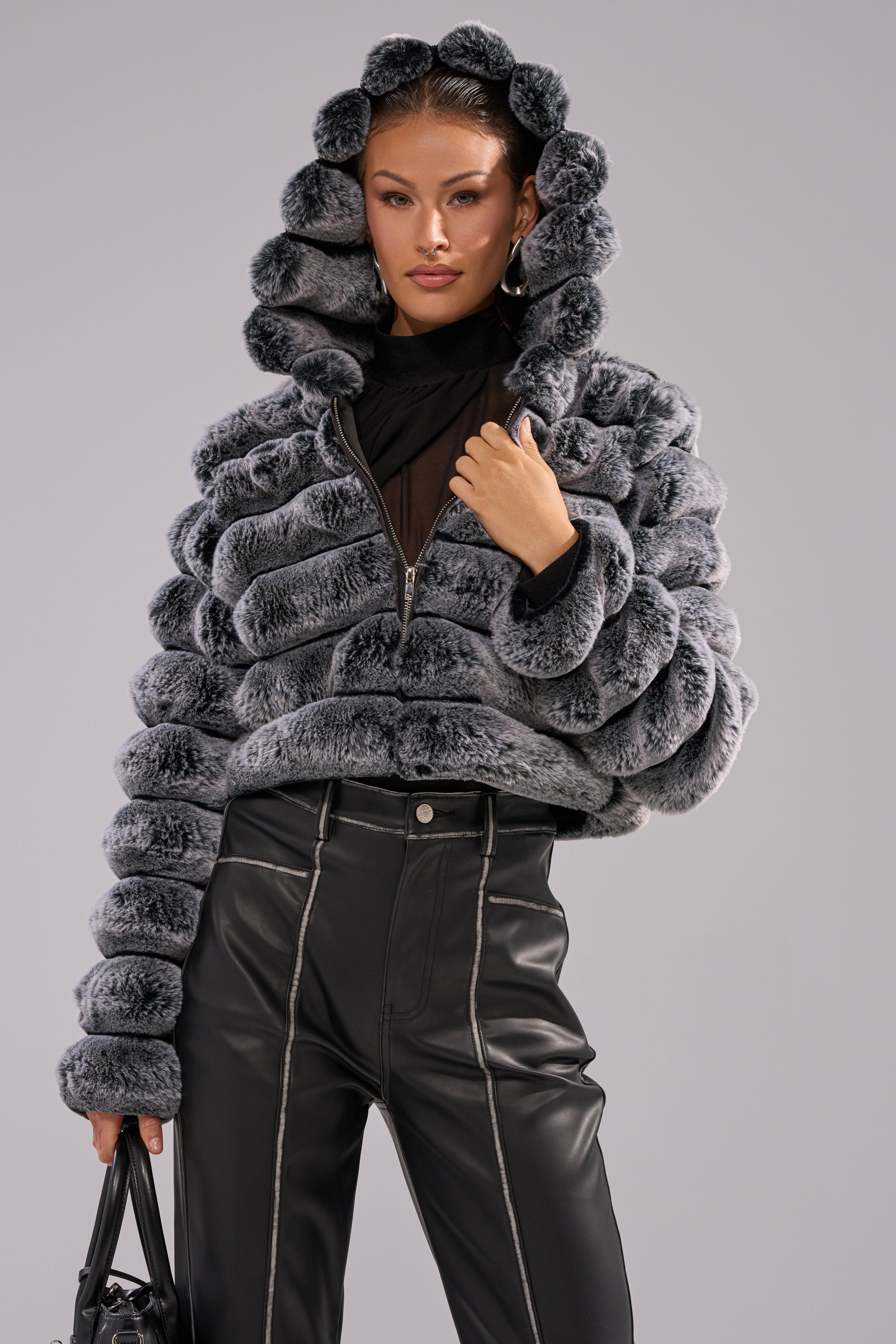 A woman poses confidently against a plain grey background, wearing the POPCORN FAUX FUR IN GREY jacket with a hood, high-waisted black leather pants, and holding a black handbag. 3