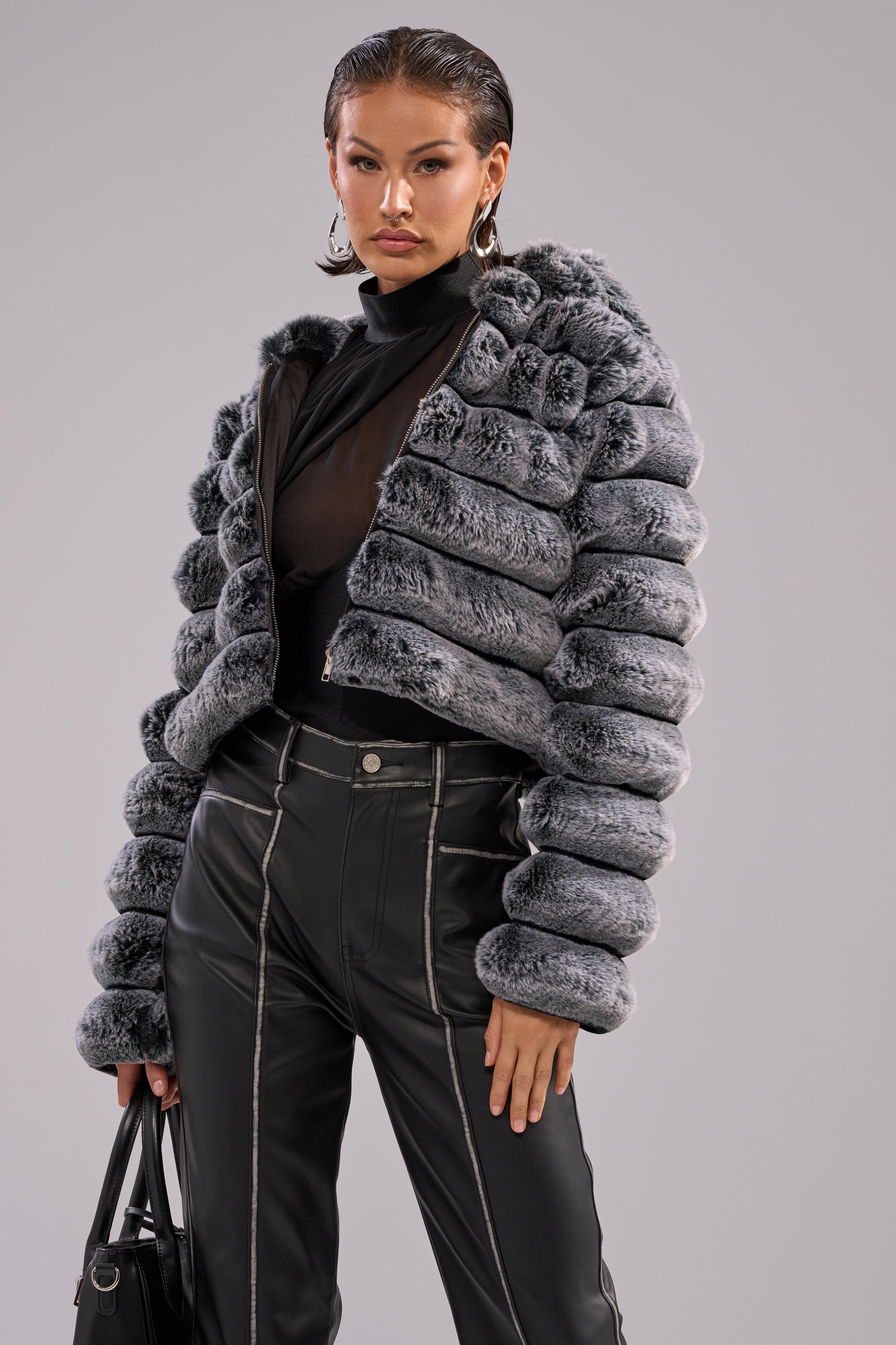 A woman stands confidently against a plain gray background, wearing the POPCORN FAUX FUR IN GREY jacket, a black sheer top, and high-waisted black leather pants with white stitching, accessorized with a black handbag. 4