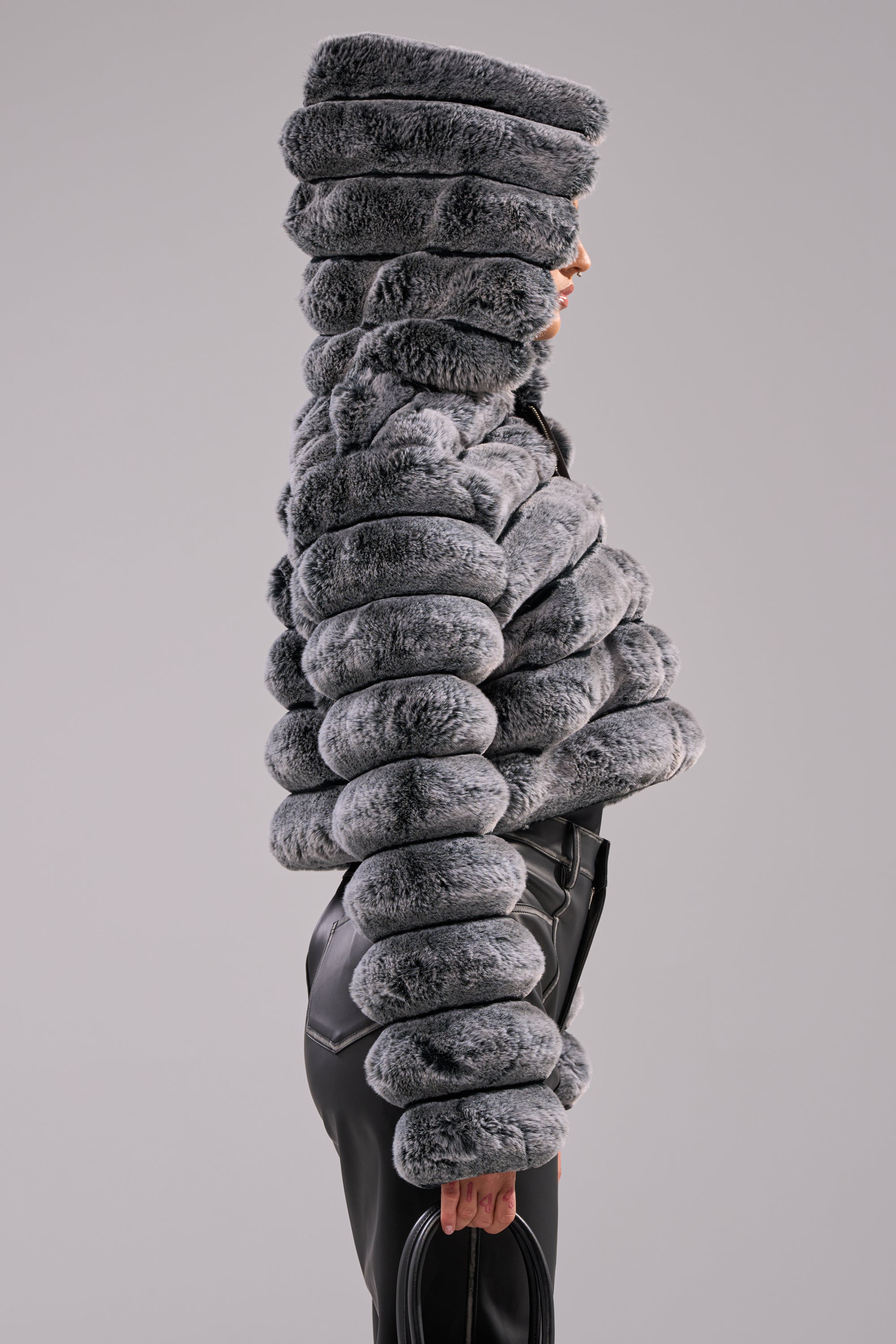 A person stands in profile against a plain background, wearing the POPCORN FAUX FUR IN GREY jacket. The large, rounded segments and cocoon-like design cover the head and arms for a sculptural, dramatic silhouette. 5
