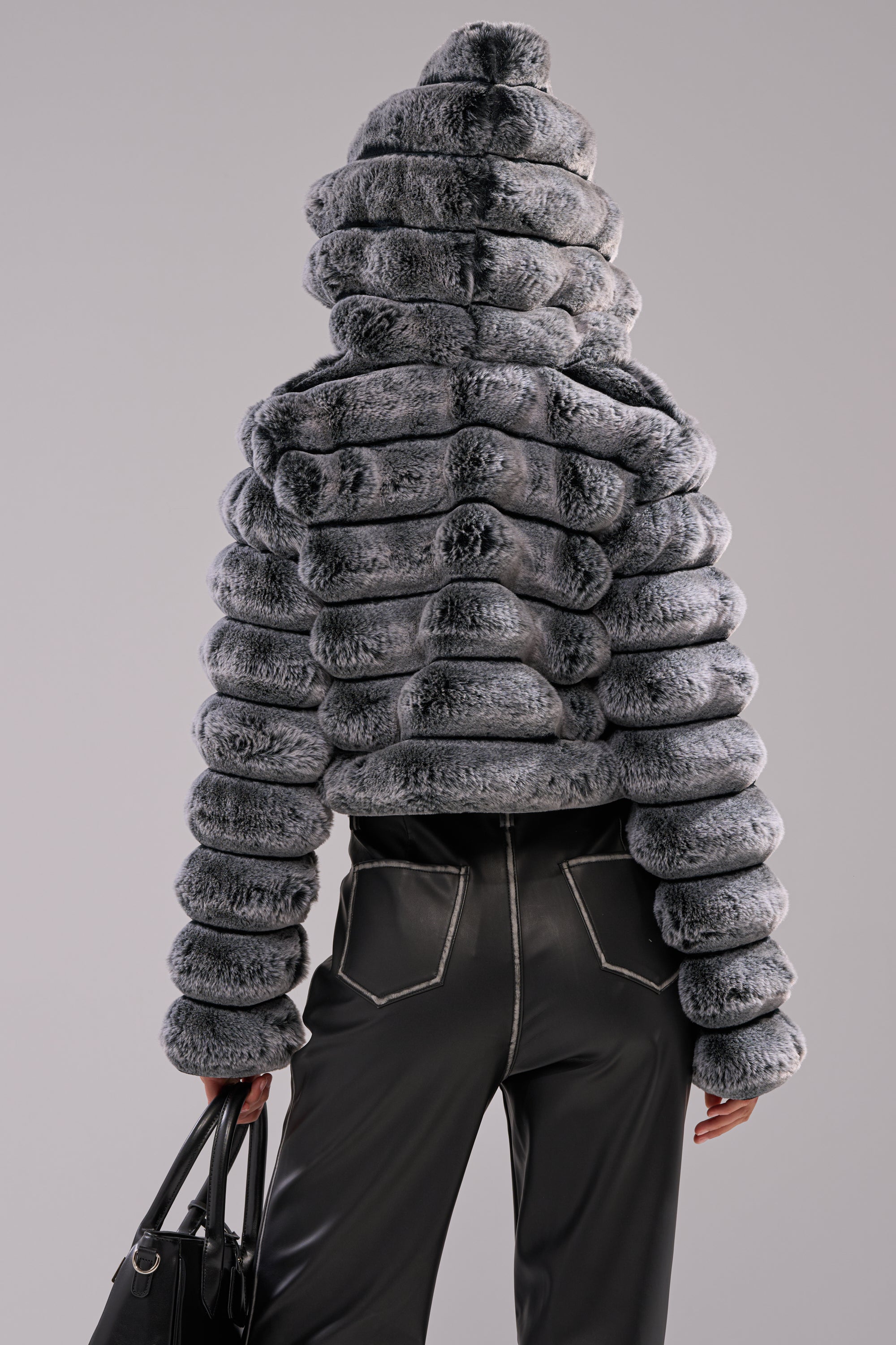 A person stands against a plain light background, wearing the POPCORN FAUX FUR IN GREY jacket with a hood and black pants. Facing away, they hold a black handbag in their left hand. 6