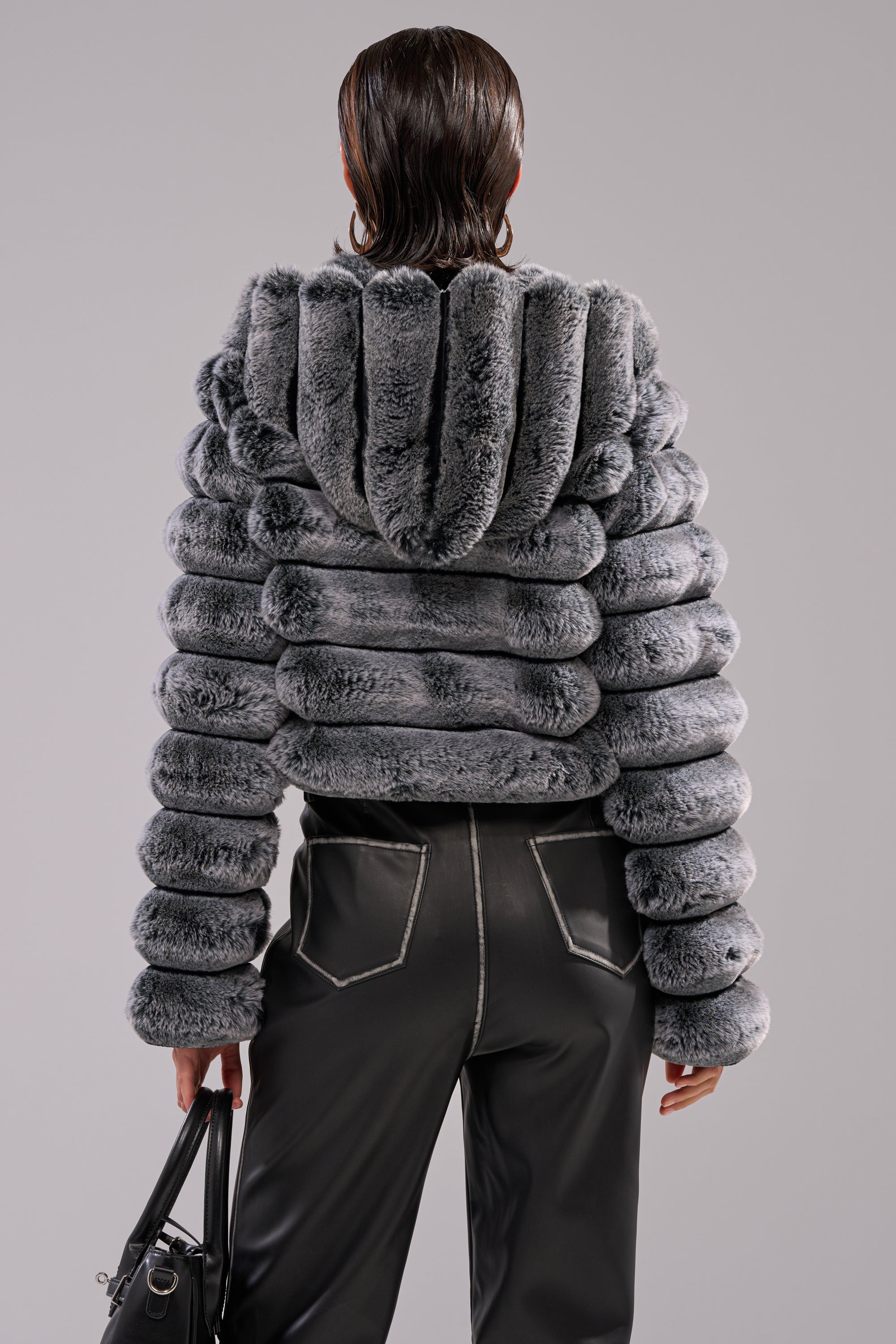 A person with slicked-back hair wears the POPCORN FAUX FUR IN GREY jacket, which is cropped with a puffy hood, paired with black pants featuring white stitching, and holds a black handbag by their side. 7