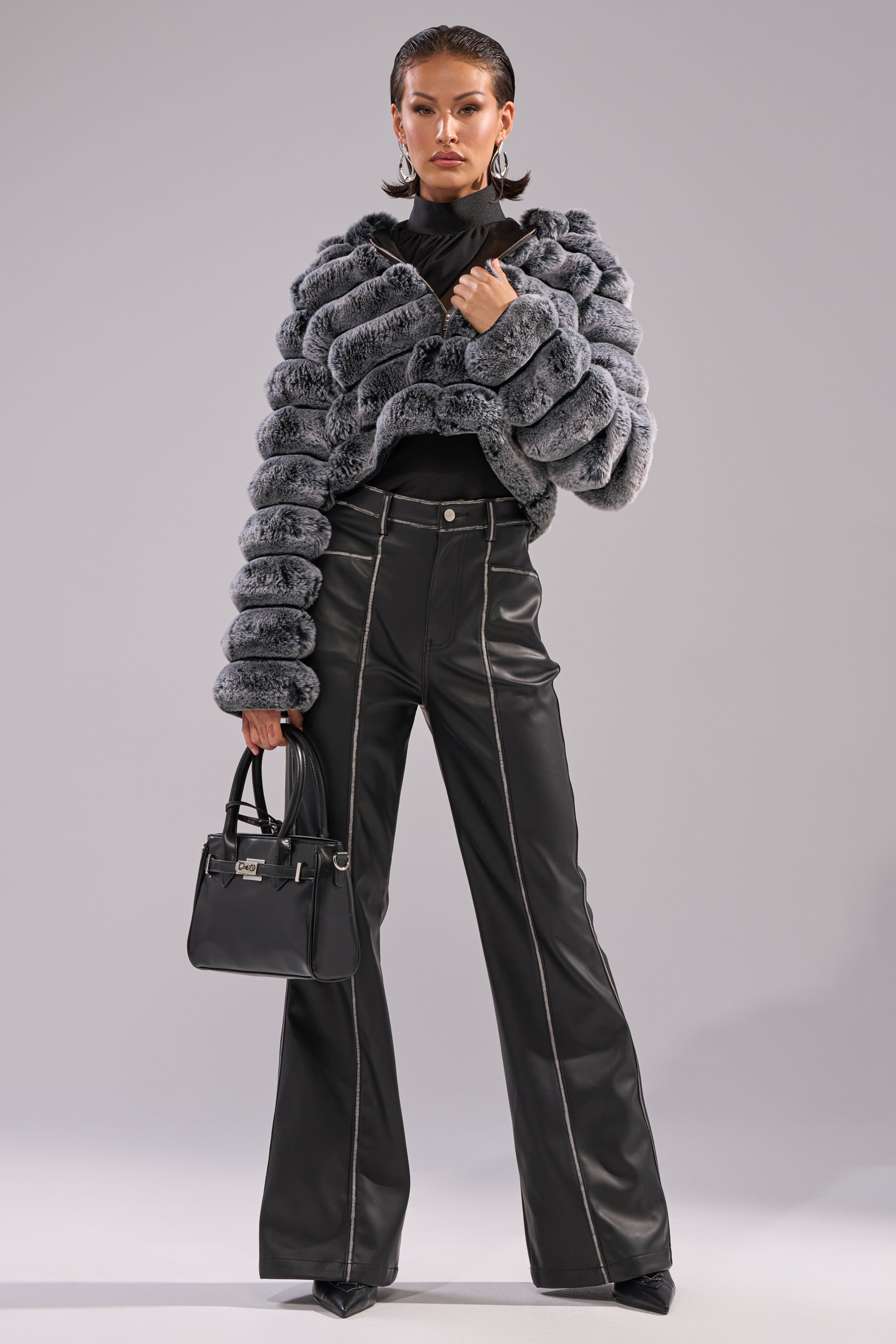 A woman poses confidently in the POPCORN FAUX FUR IN GREY jacket, paired with black high-waisted wide-leg pants with white stitching, pointed black heels, a small black handbag, statement earrings, and slicked-back hair. 8