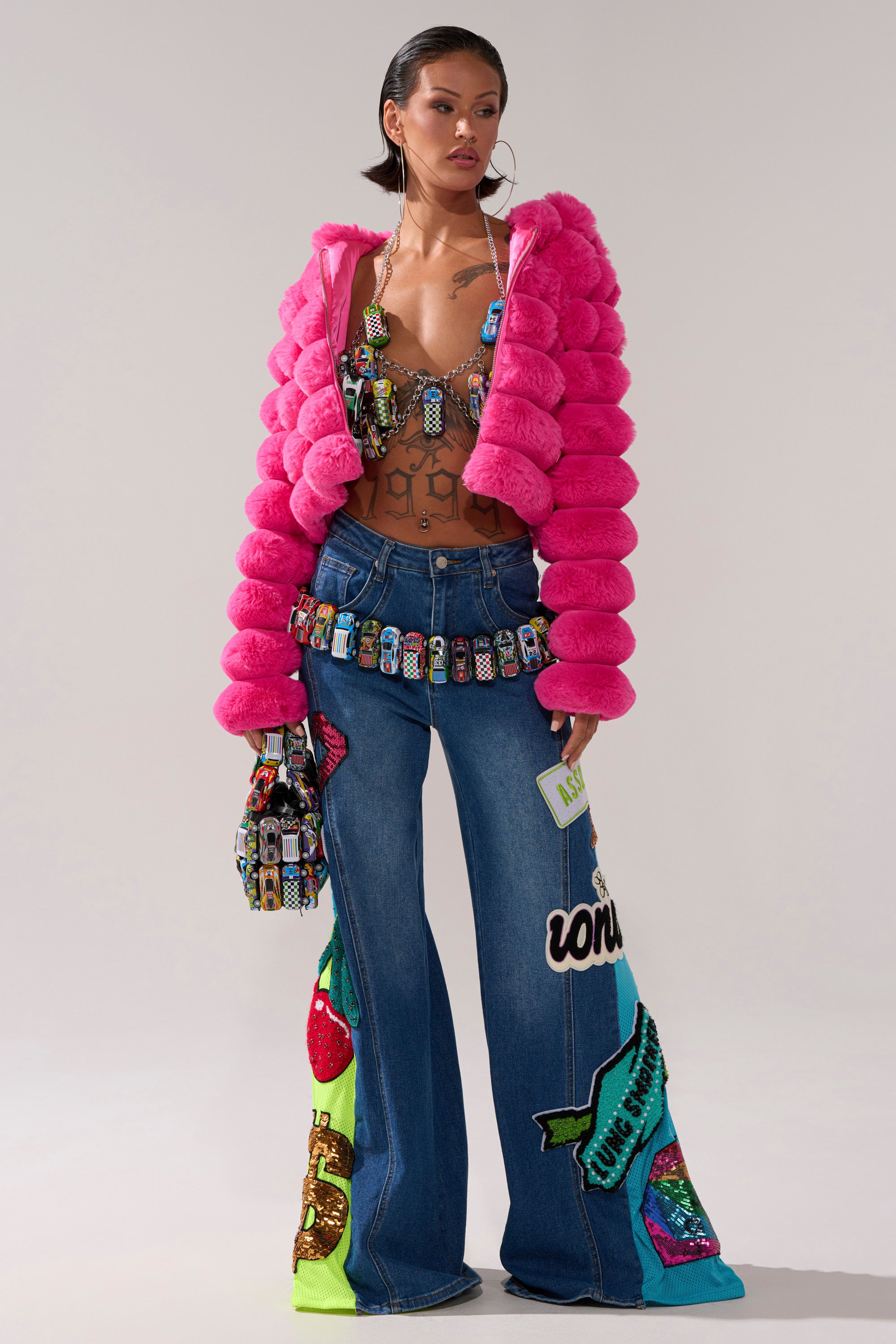 A woman poses in the POPCORN FAUX FUR IN PINK jacket, paired with a beaded bra, wide-leg jeans with colorful appliqué, a jeweled belt, and a patterned handbag against a neutral background.
