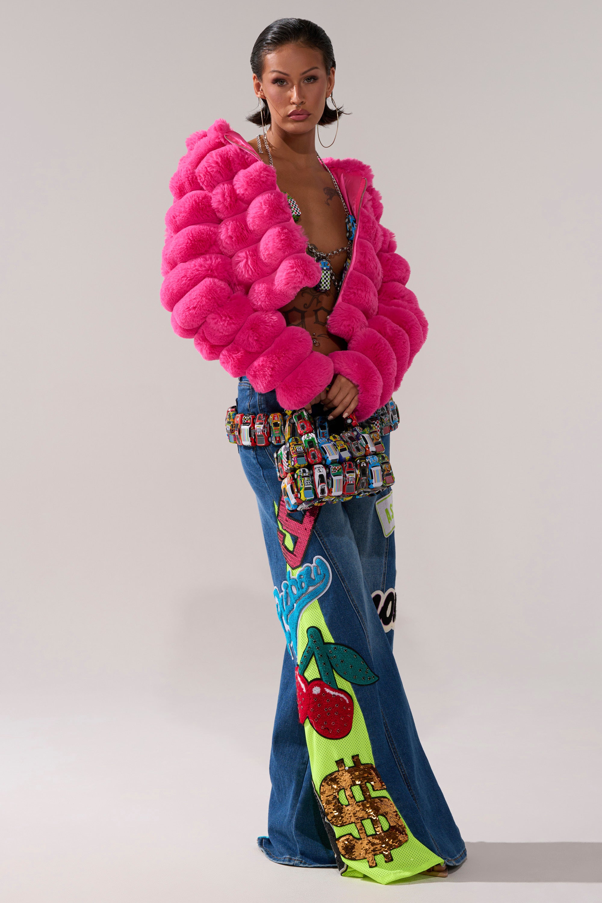 A woman wears the POPCORN FAUX FUR IN PINK jacket, a colorful patterned crop top, and wide-leg blue jeans with vivid cherry and waffle graphics. She carries a multicolored handbag.