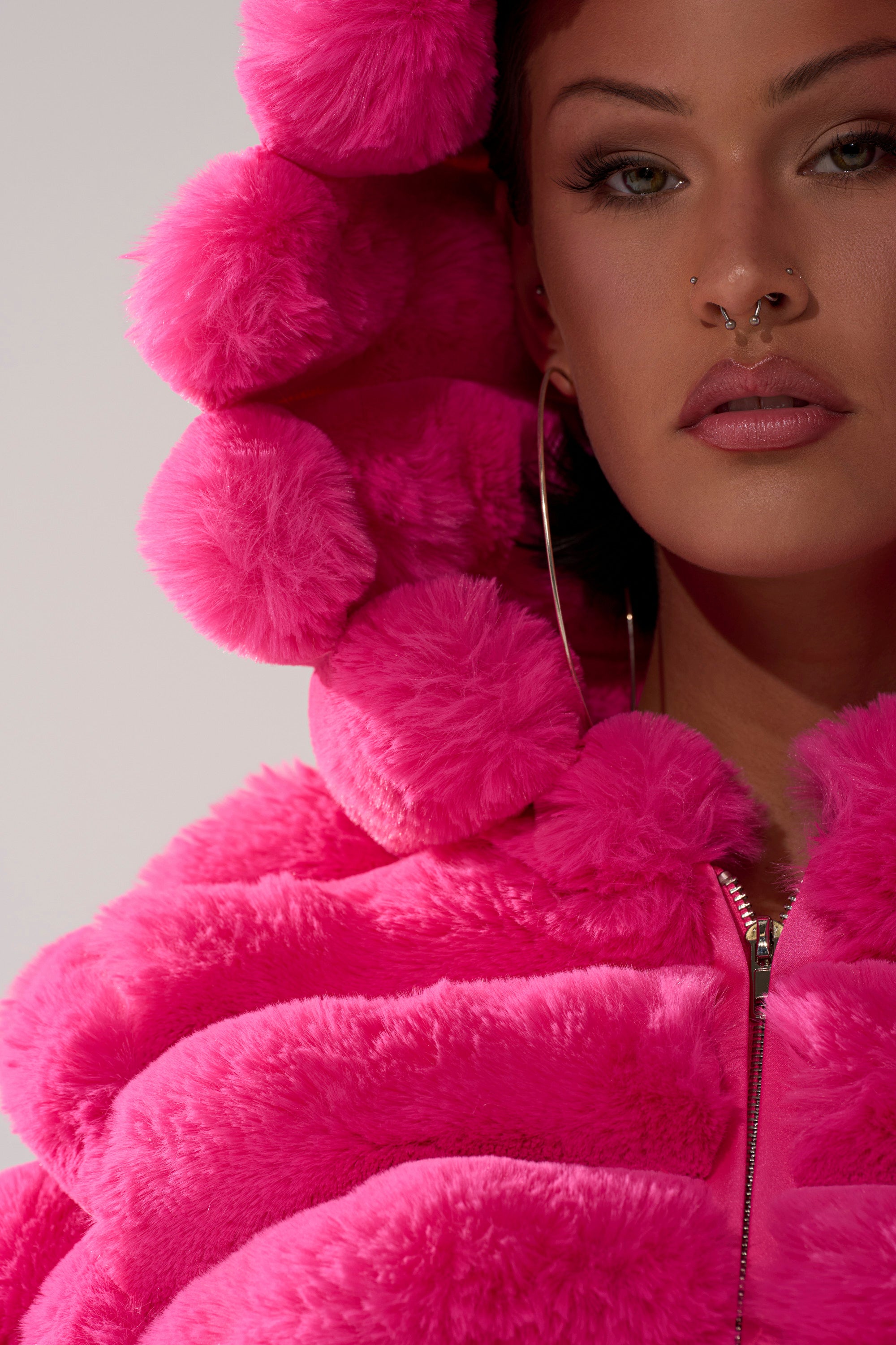 Wearing the POPCORN FAUX FUR IN PINK jacket with oversized round pom-poms on the hood, a person shows only their lower face and upper torso. They have a septum piercing and wear large hoop earrings.