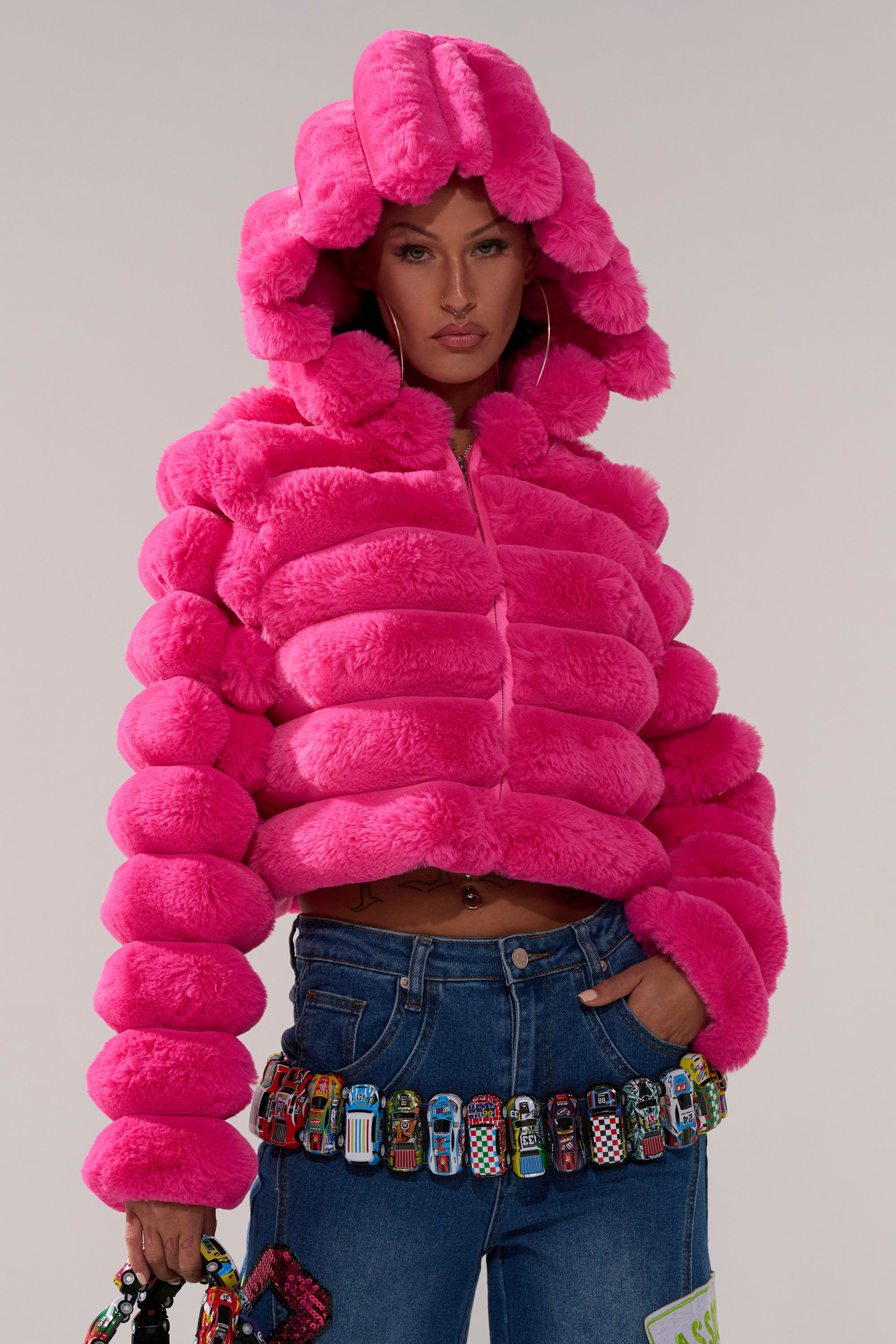 A person wears the POPCORN FAUX FUR IN PINK jacket with rounded sections, paired with blue jeans and a matching bag decorated with toy cars. The background is plain and neutral.