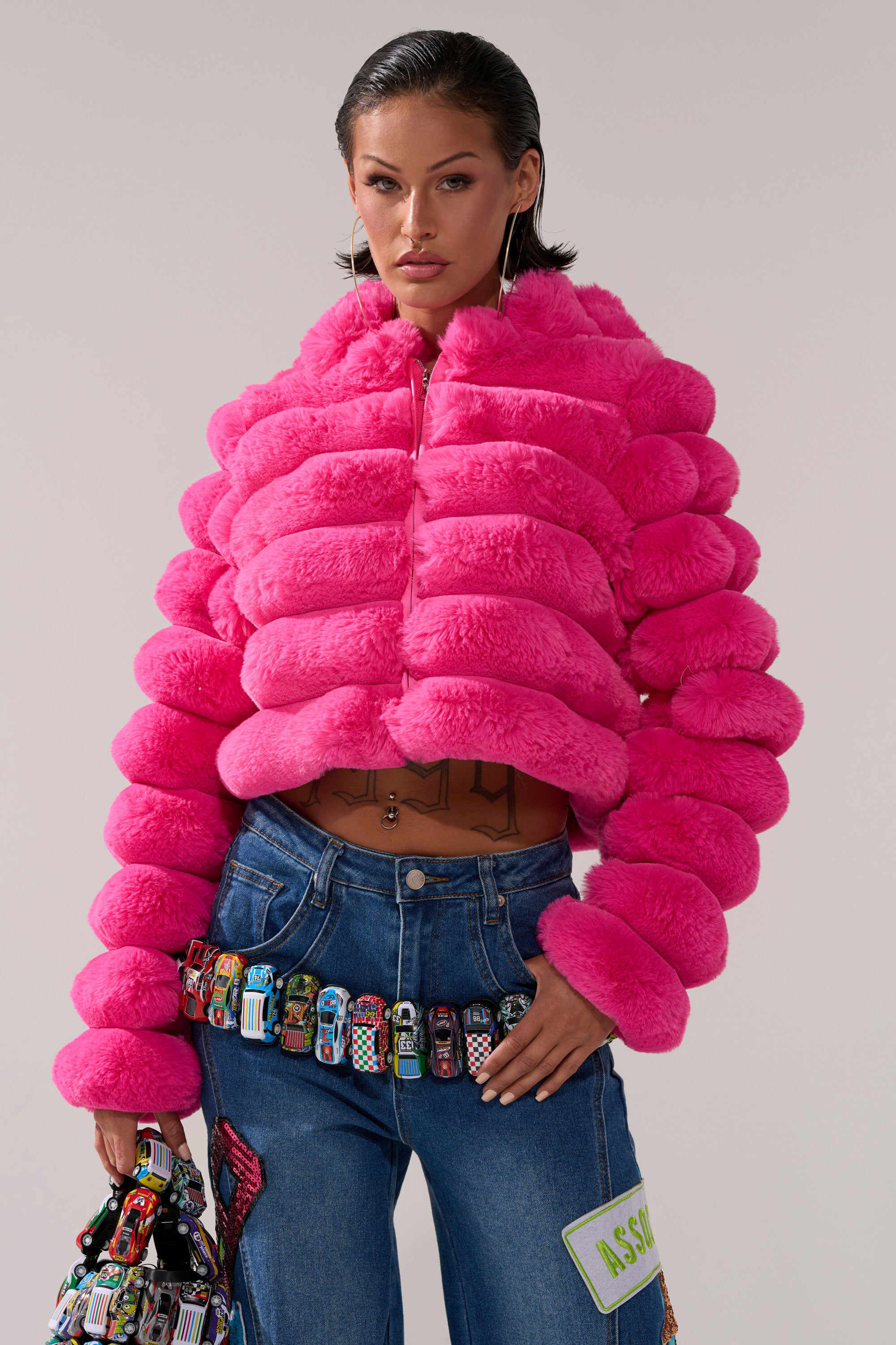 A woman stands facing forward in the POPCORN FAUX FUR IN PINK jacket and blue jeans with colorful toy cars, holding a matching toy car bag against a plain background.