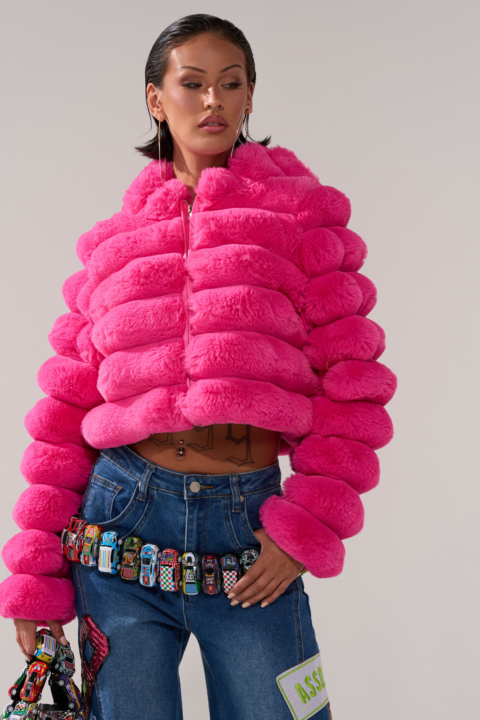 Wearing the POPCORN FAUX FUR IN PINK jacket and blue jeans adorned with colorful toy cars and patches, a person holds a matching accessory against a plain light background, adding bold flair to the playful look.