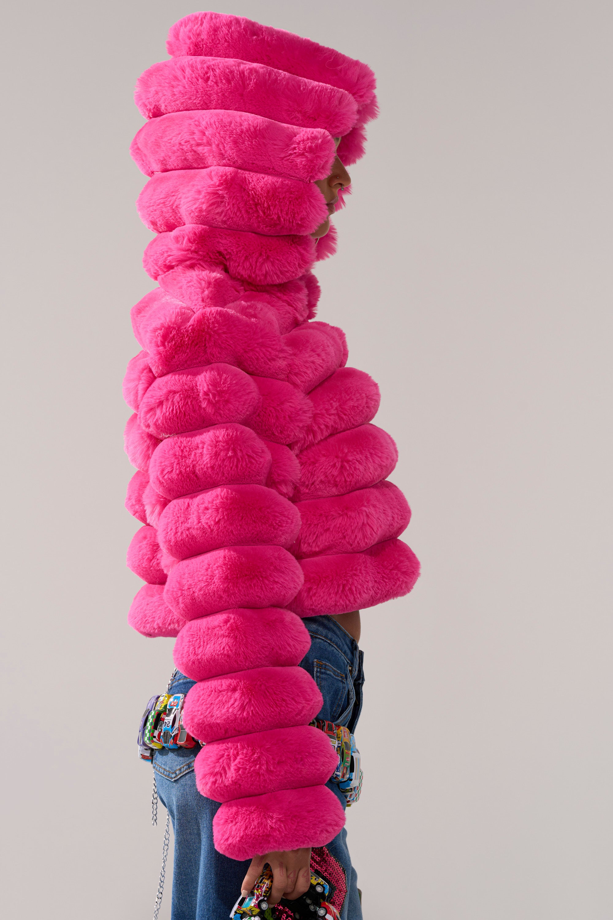 A person in profile wears the POPCORN FAUX FUR IN PINK jacket with a large hood, styled with blue jeans and a colorful beaded purse, standing against a plain background.