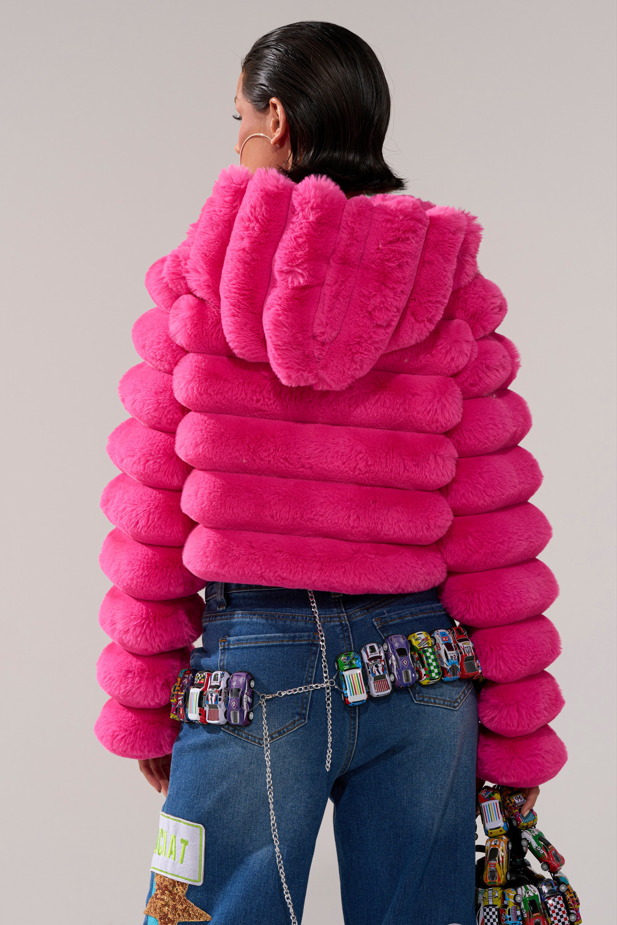 Seen from behind, a person with slicked-back hair wears the POPCORN FAUX FUR IN PINK jacket—a hooded, bright pink cropped piece ideal for winter—styled with blue jeans adorned with colorful chains, patches, and small toy cars.