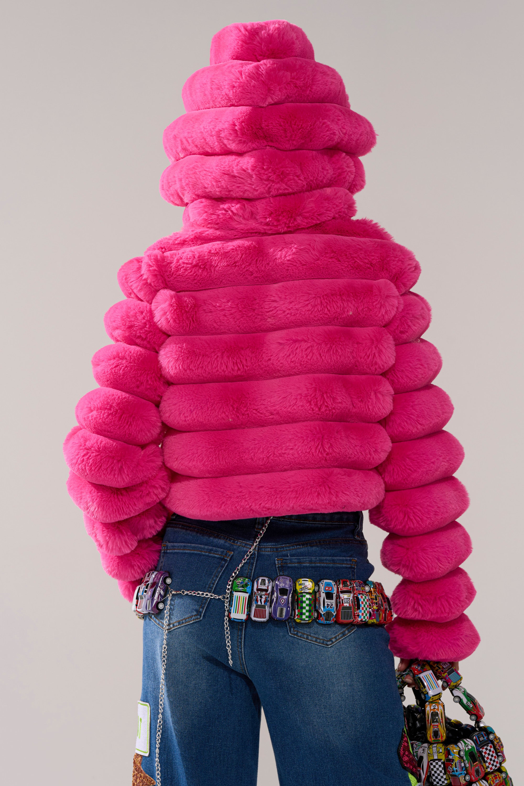 Wearing the POPCORN FAUX FUR IN PINK jacket and blue jeans adorned with colorful toy cars, a person stands with their back to the camera against a plain background.