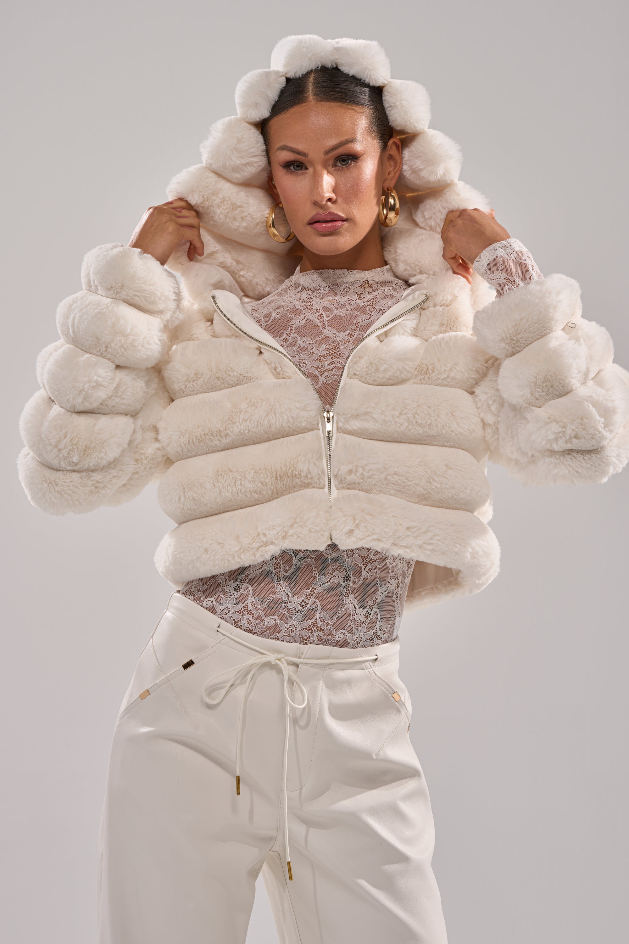 A woman models the POPCORN FAUX FUR IN WHITE—a cropped white faux fur jacket with a hood—over a lace top and high-waisted pants, holding the hood as she looks confidently at the camera against a plain backdrop. 0