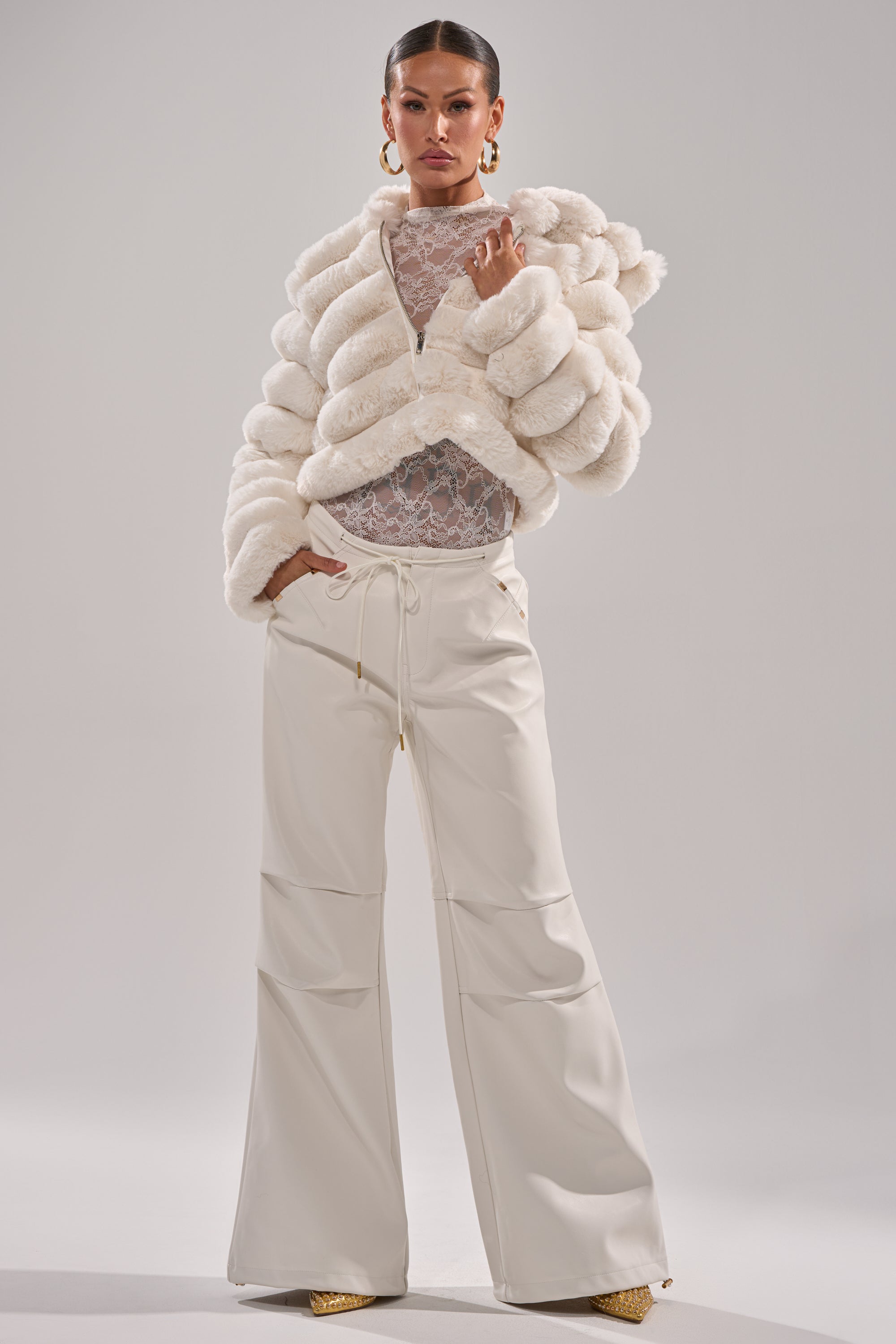 A woman poses confidently against a plain light background in the POPCORN FAUX FUR IN WHITE jacket, paired with a sheer lace top, wide-leg cream trousers, large hoop earrings, and gold shoes. 9