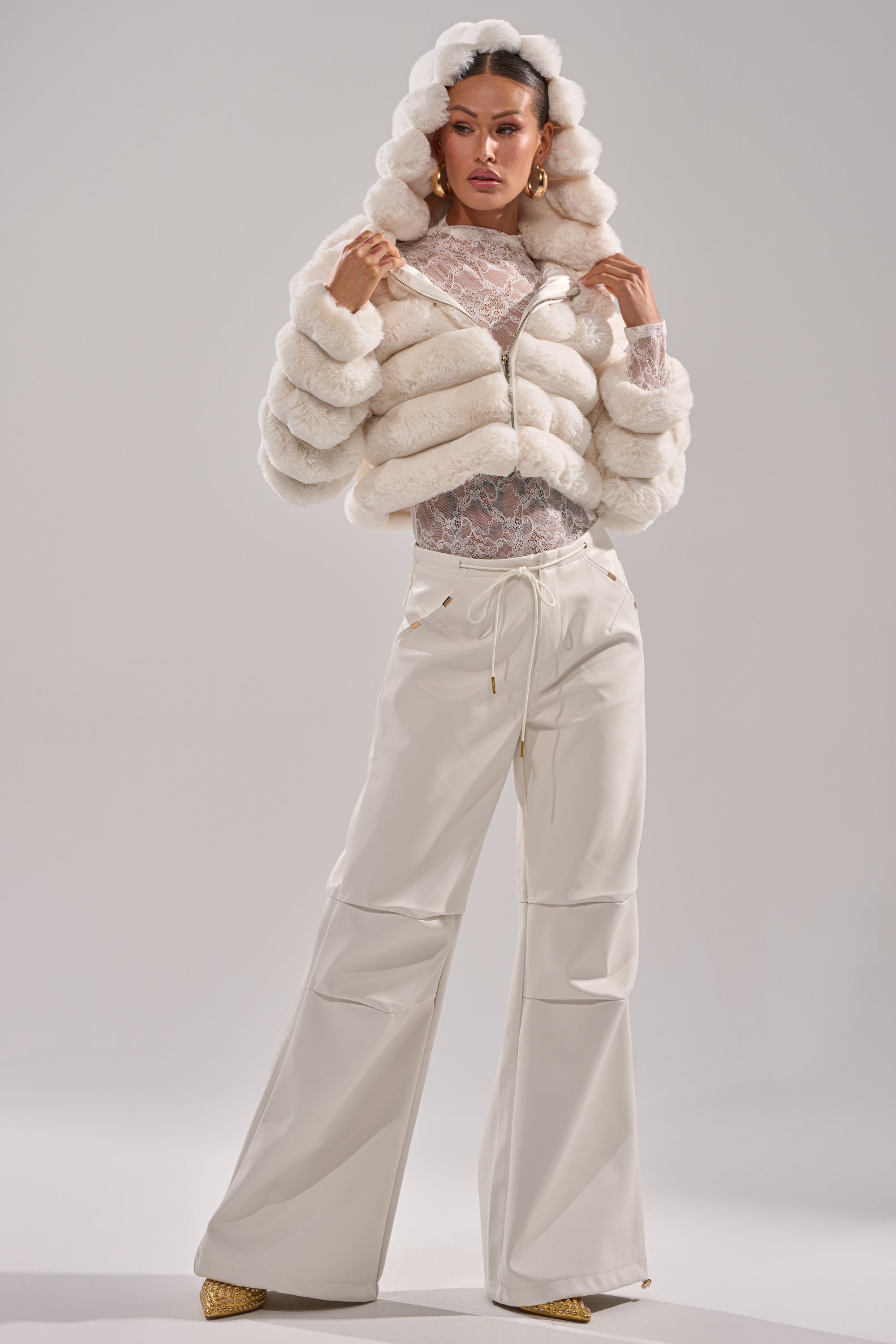 A woman confidently models the POPCORN FAUX FUR IN WHITE jacket over a sheer lace top, styled with wide-leg cream trousers and gold heels, capturing chic winter fashion against a plain light background. 10
