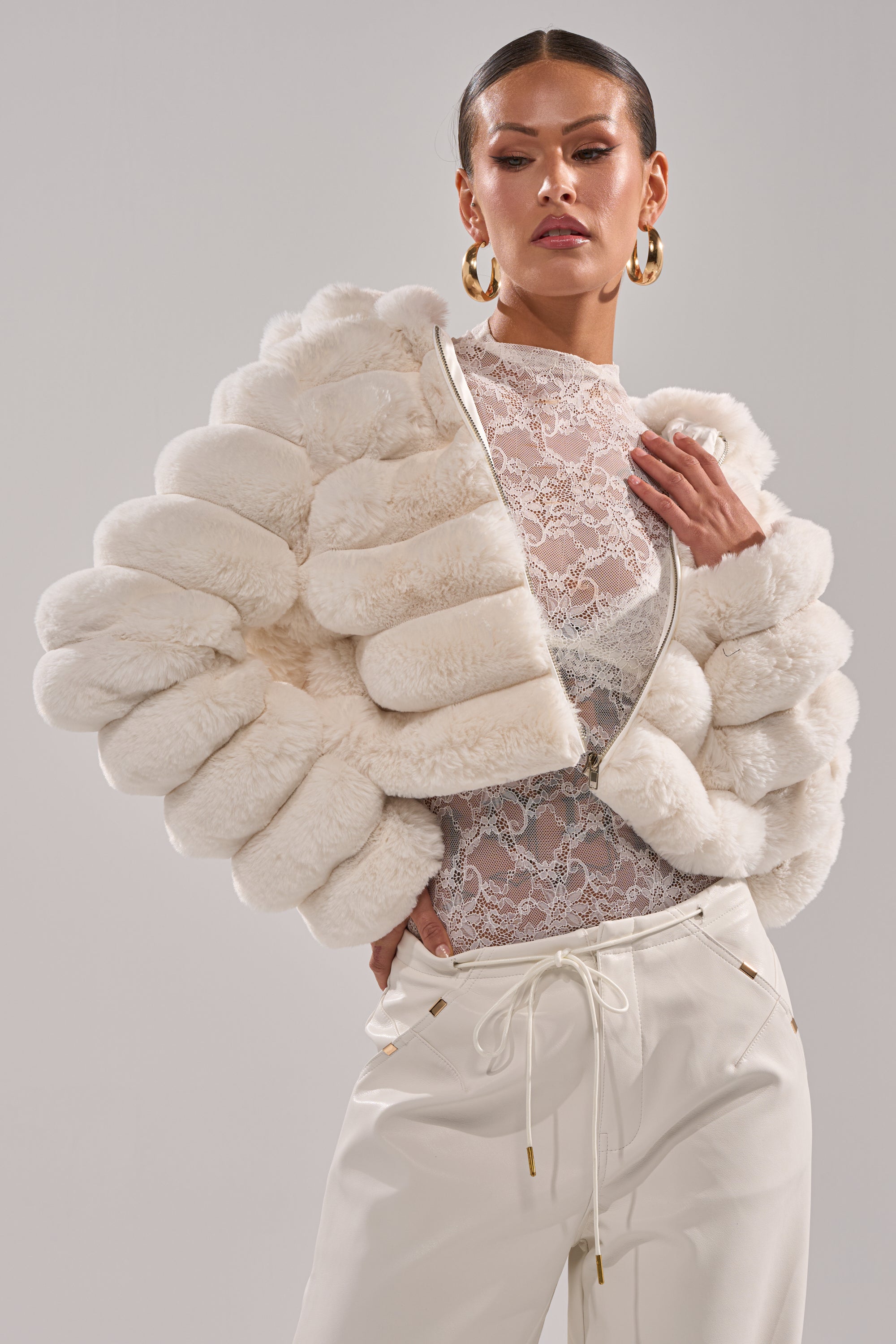 A woman poses confidently in the POPCORN FAUX FUR IN WHITE, styled over a sheer lace top and white pants with gold accents, accessorized with bold makeup, slicked-back hair, and large gold hoop earrings. 1
