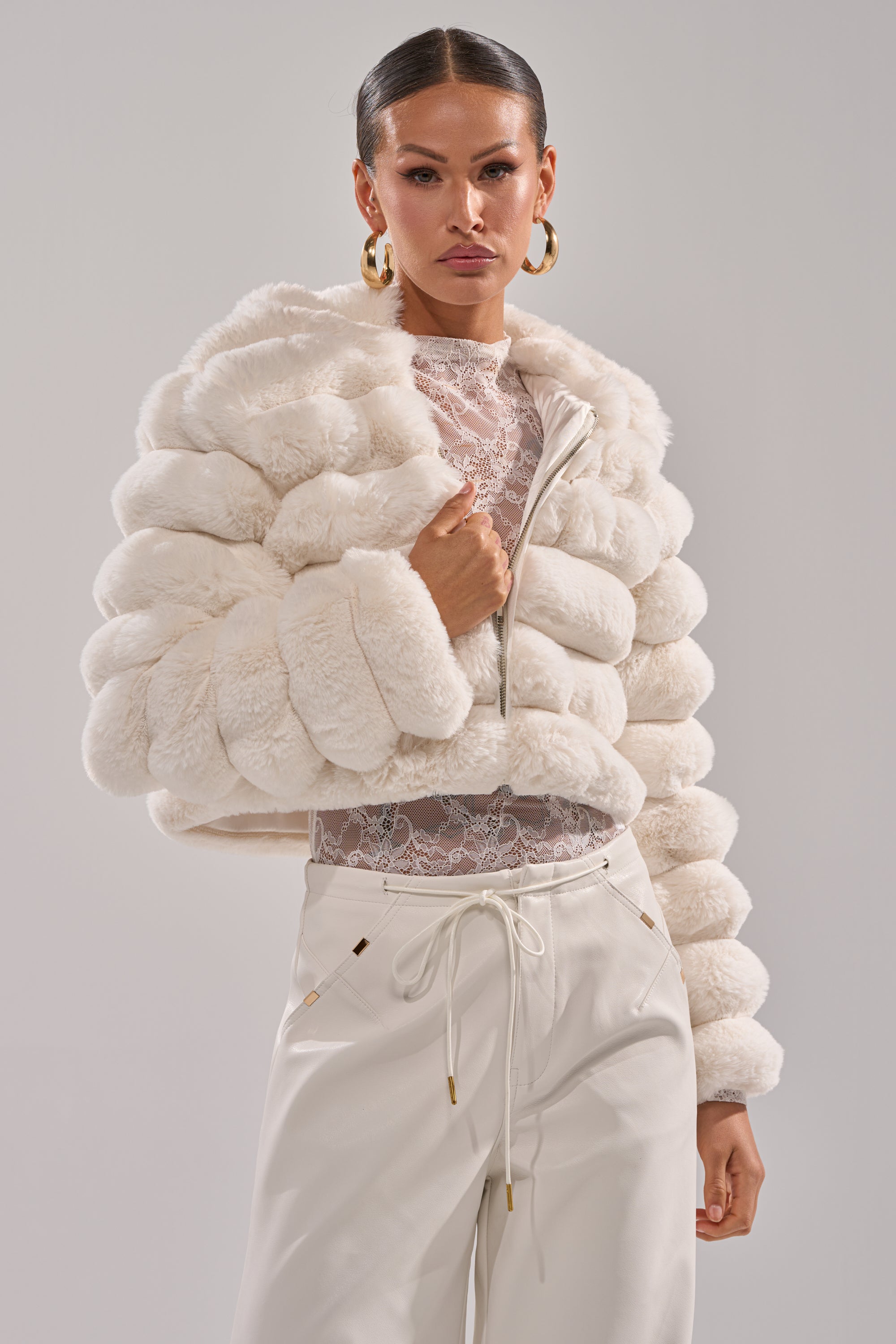 A woman poses confidently in the POPCORN FAUX FUR IN WHITE over a sheer lace top and high-waisted pants, her slicked-back hair and large gold hoop earrings enhancing her chic style against a plain light background. 2