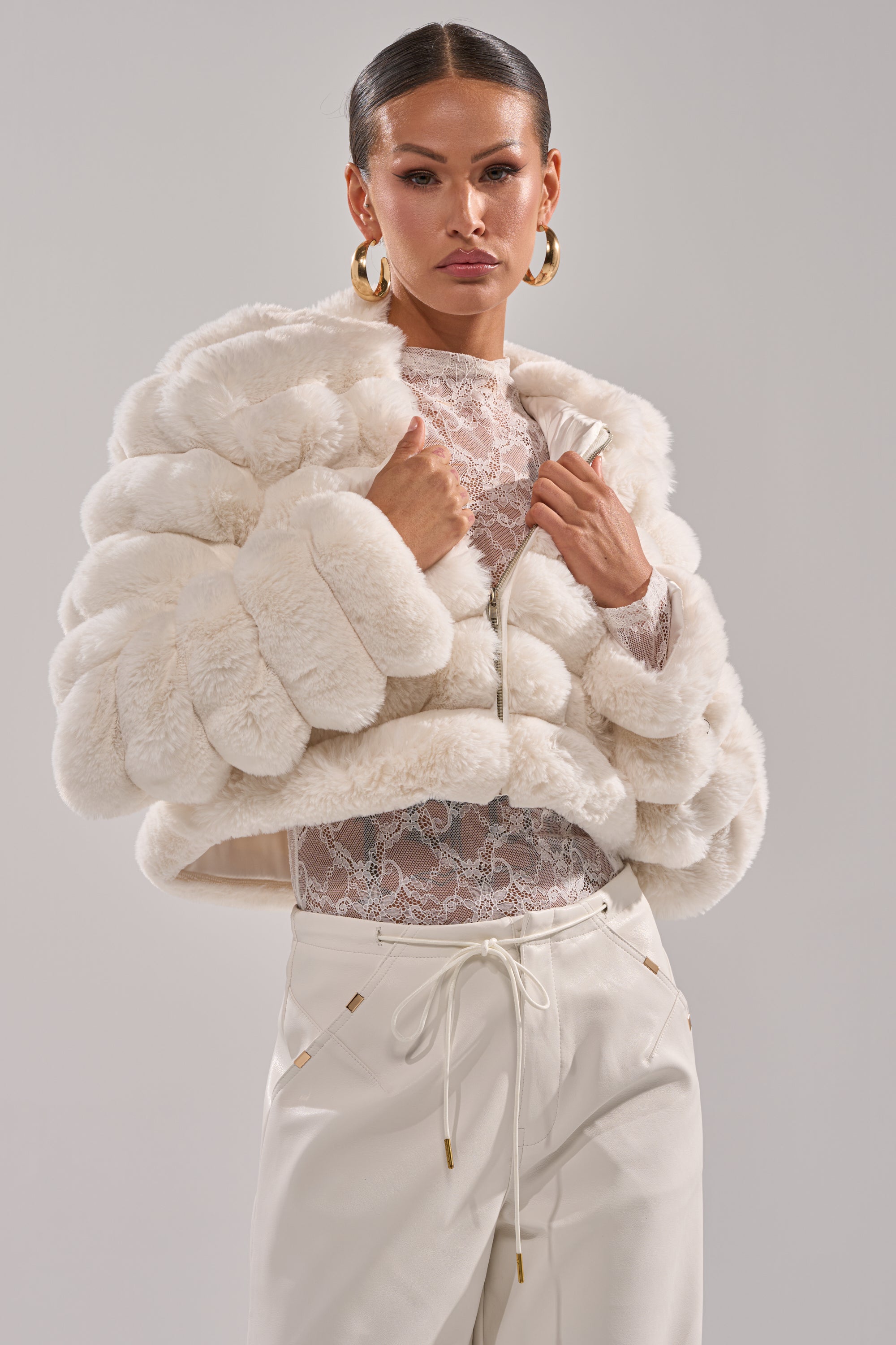 A woman wears the POPCORN FAUX FUR IN WHITE jacket over a sheer lace top and high-waisted white pants with gold accents, posing confidently against a plain light gray background. 3