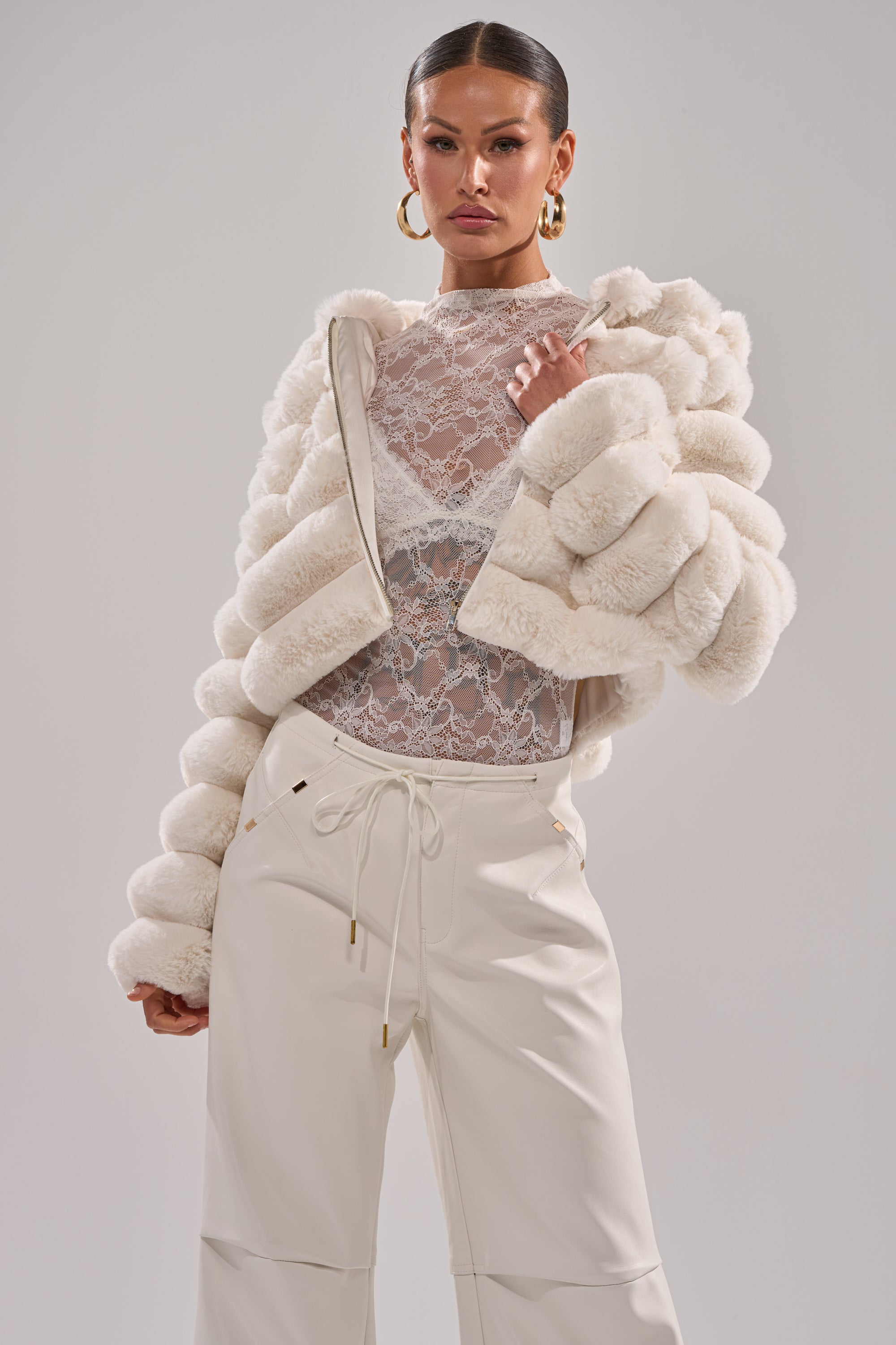 A woman stands confidently against a plain background in the POPCORN FAUX FUR IN WHITE, paired with a sheer lace top and high-waisted white pants. She has slicked-back hair and gold hoop earrings. 4